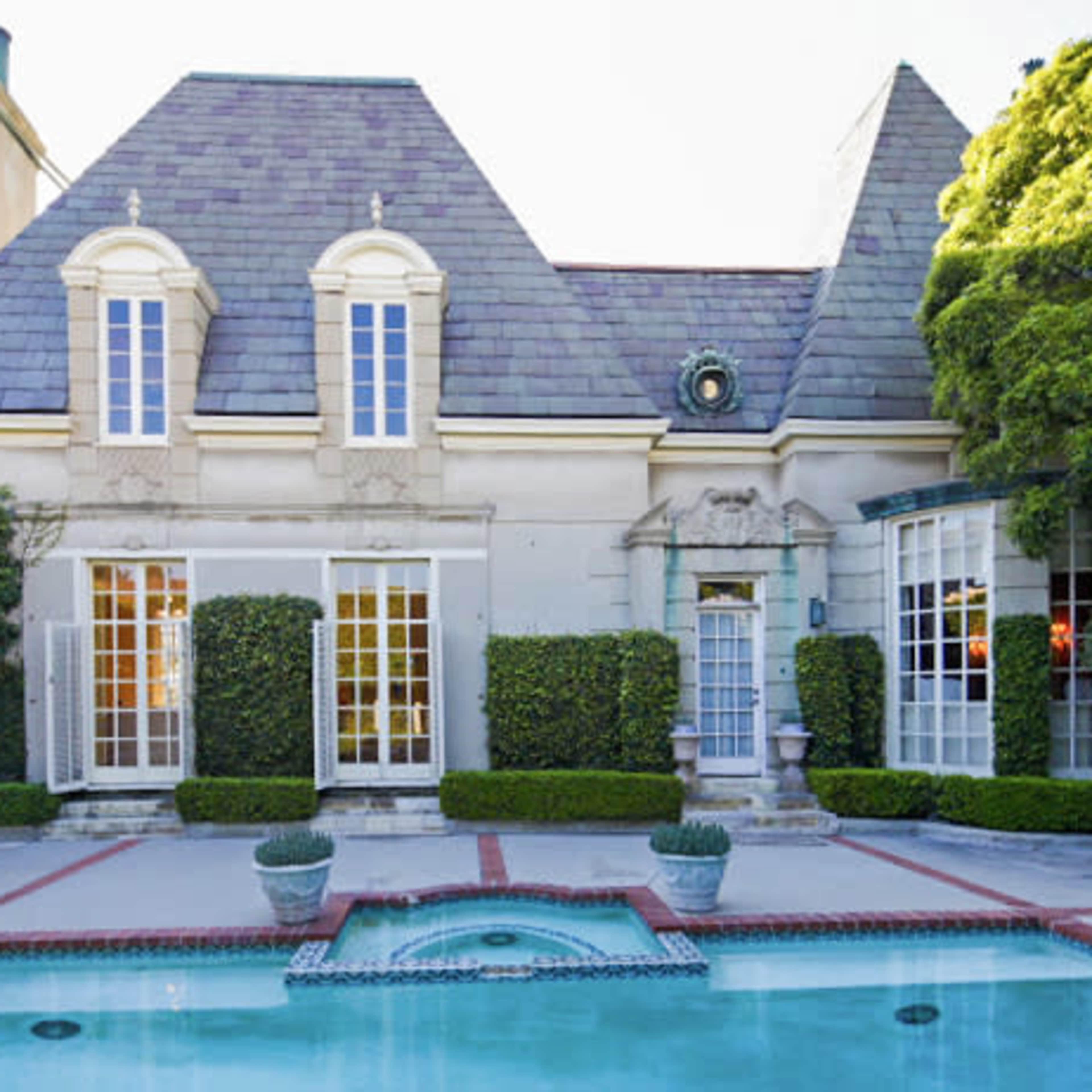 A two-story house with a slate roof is surrounded by neatly trimmed hedges and features a swimming pool in the foreground.