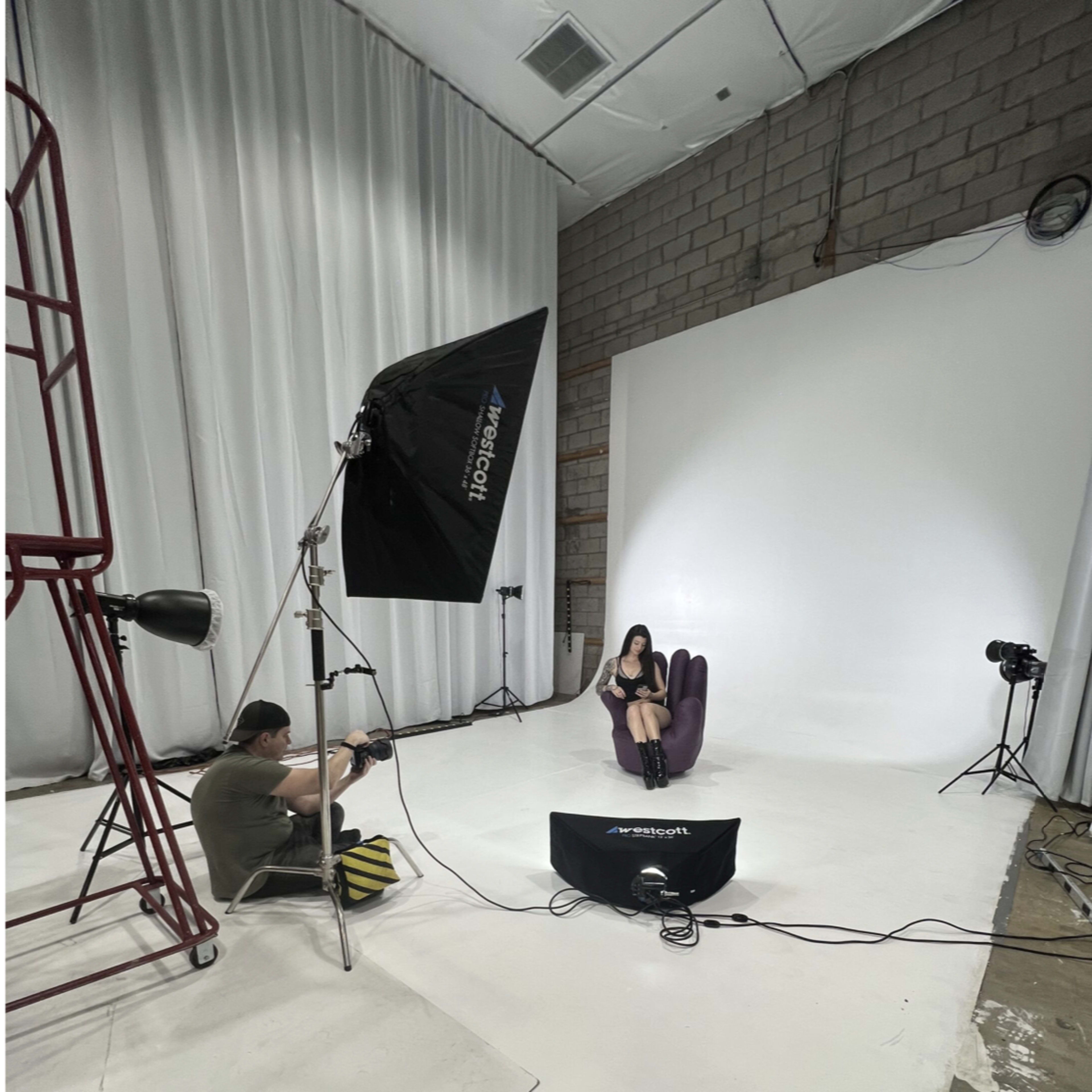 A photoshoot is taking place in a studio, featuring a model sitting on a chair while a photographer captures the scene with studio lighting equipment set up around them.