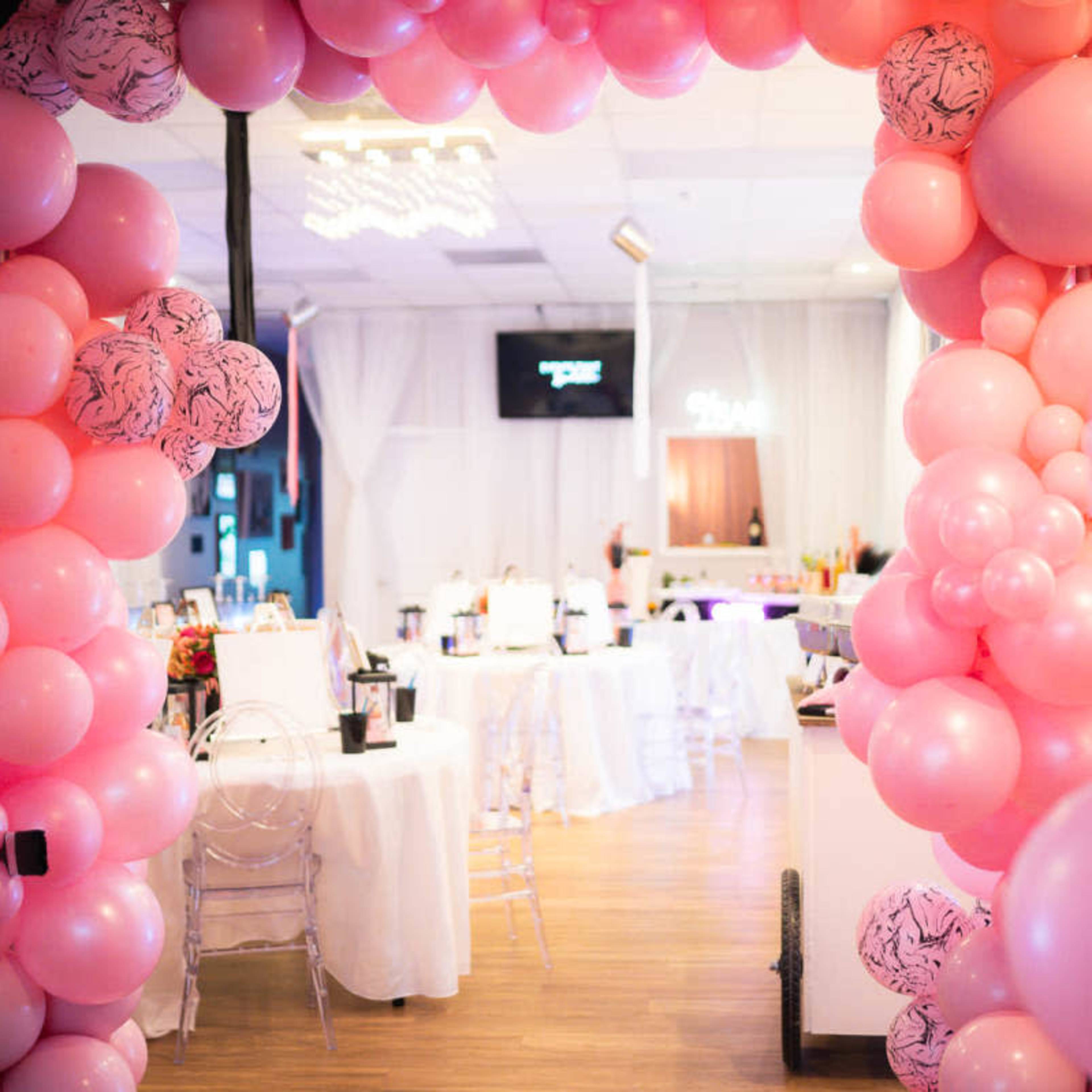 A large arch made of pink balloons frames a decorated event space featuring tables and a bar.