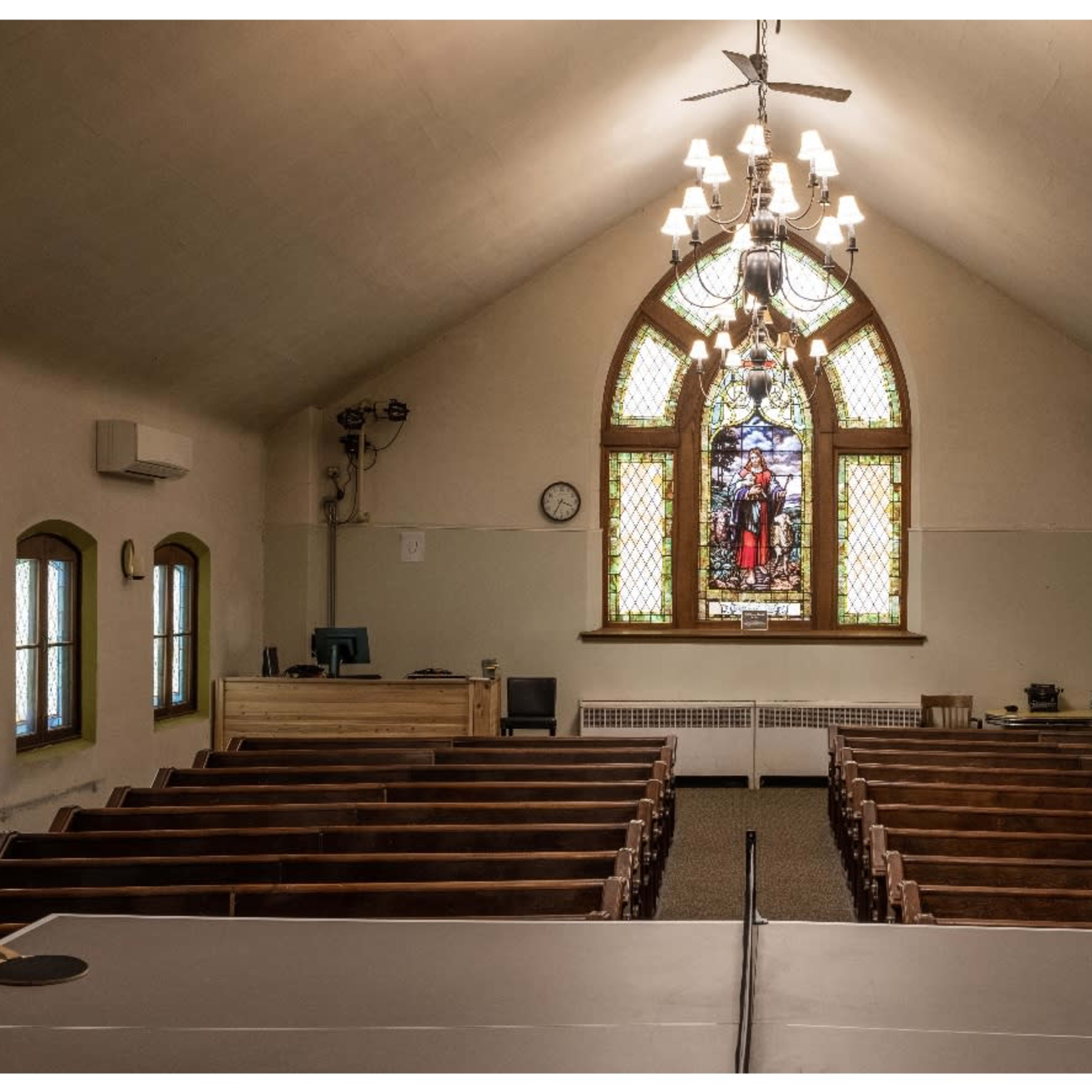 An interior view of a church, featuring wooden pews, a stained glass window, and a chandelier.