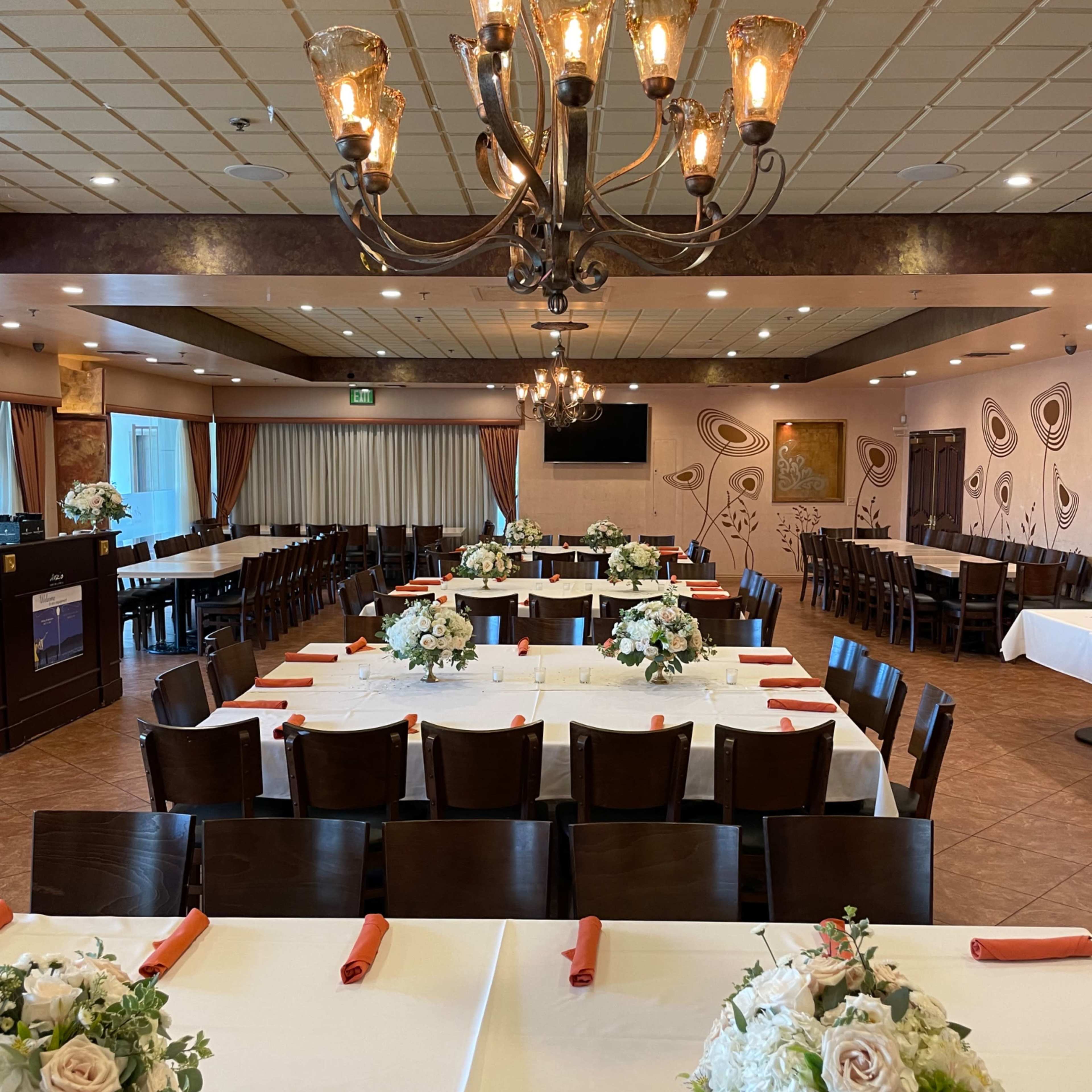 A dining room arranged with several tables set for a formal event, featuring floral centerpieces and a chandelier overhead.