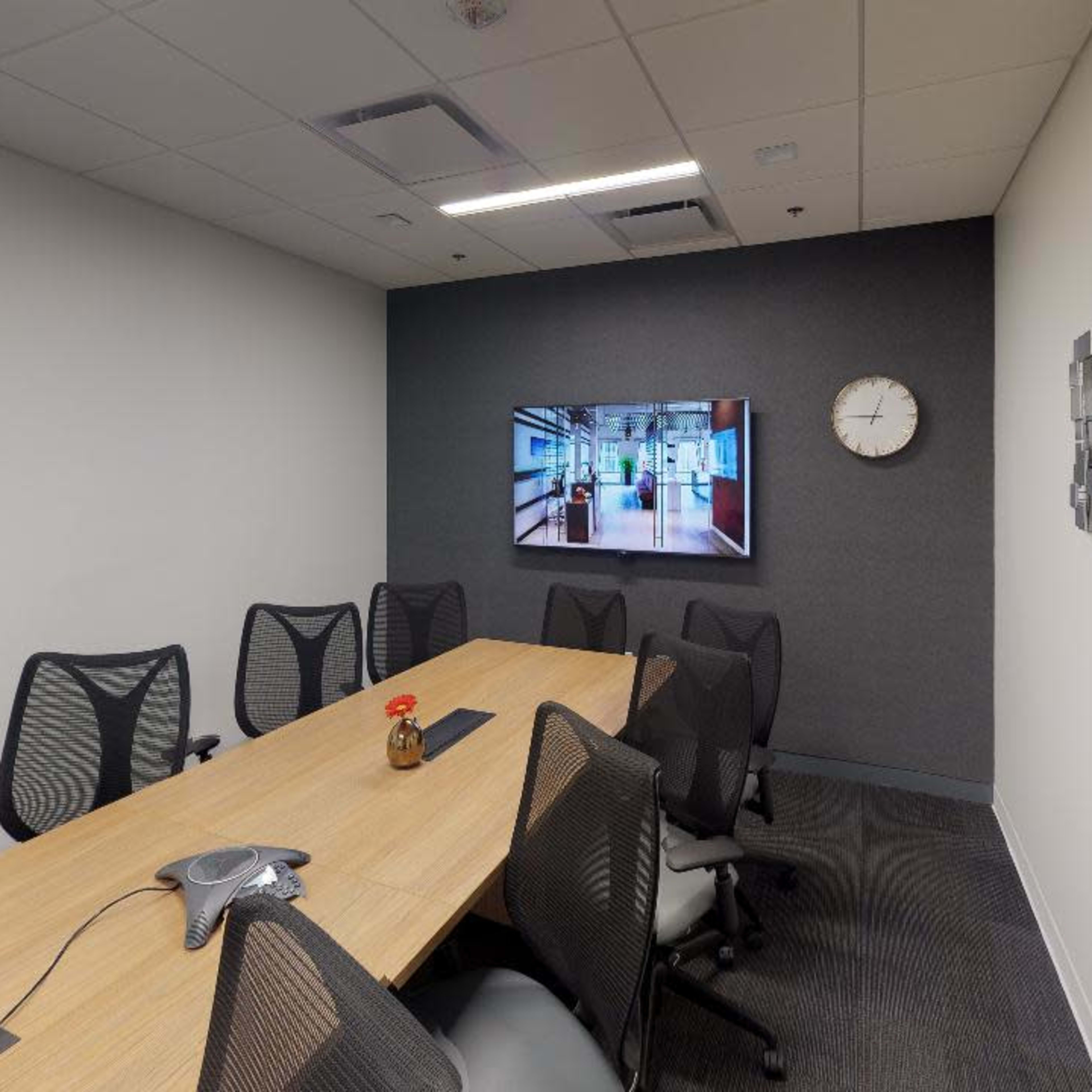 An empty conference room features a long wooden table surrounded by ergonomic chairs, a large wall-mounted screen, and a clock.
