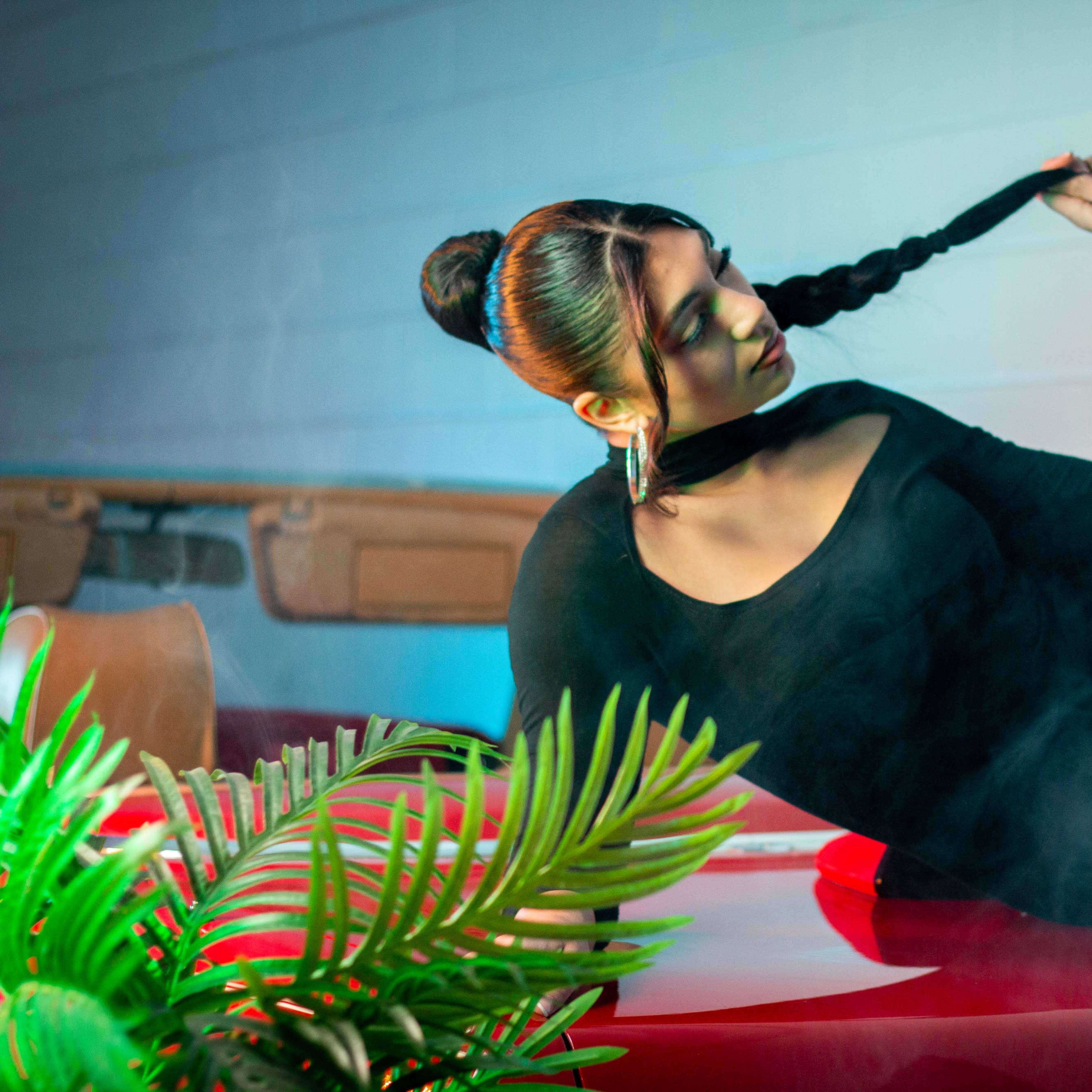 A woman in a black dress poses on the hood of a red car under a smoky atmosphere, with green plants in the foreground.