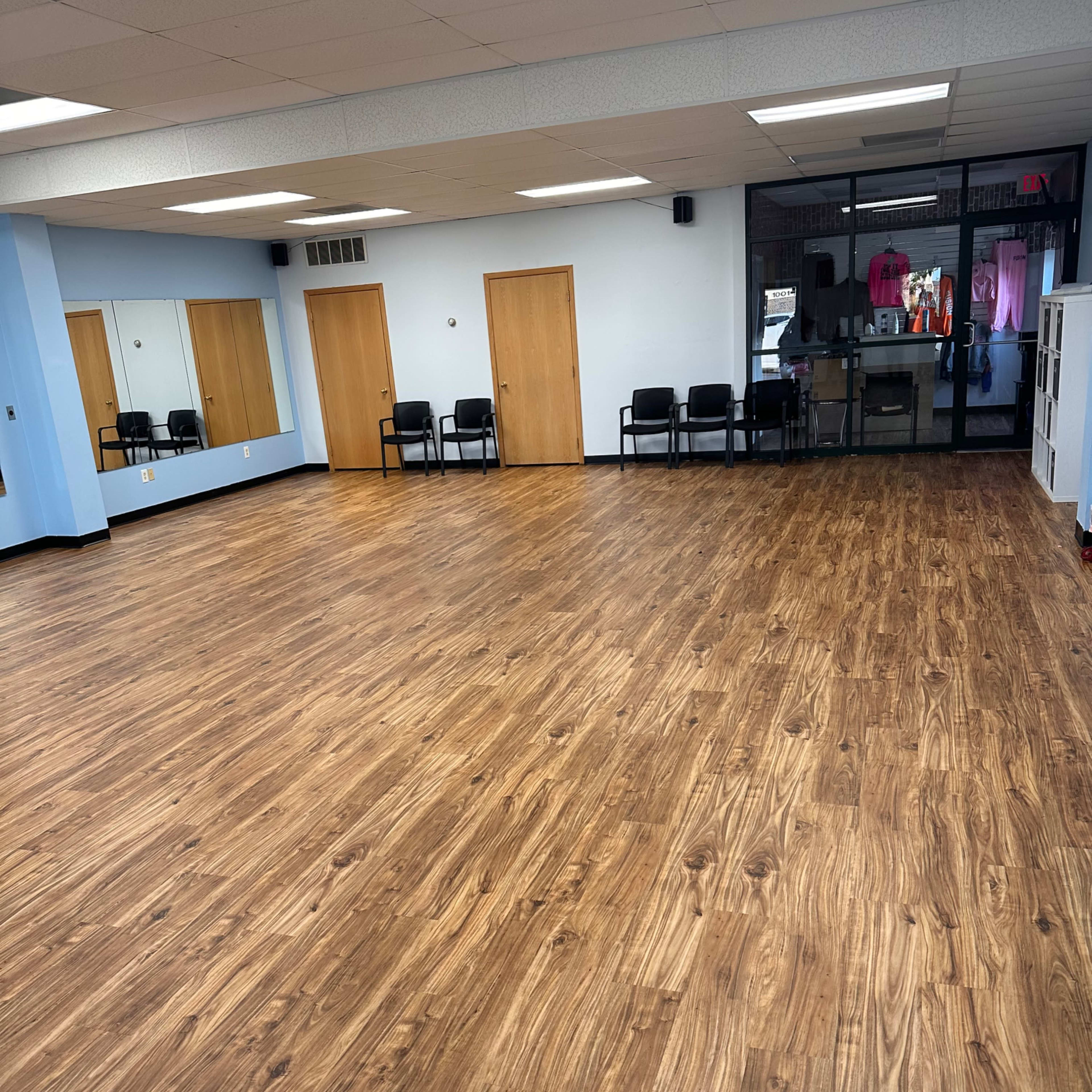 A large, empty room with wooden flooring, mirrors on one wall, and several black chairs arranged on one side.