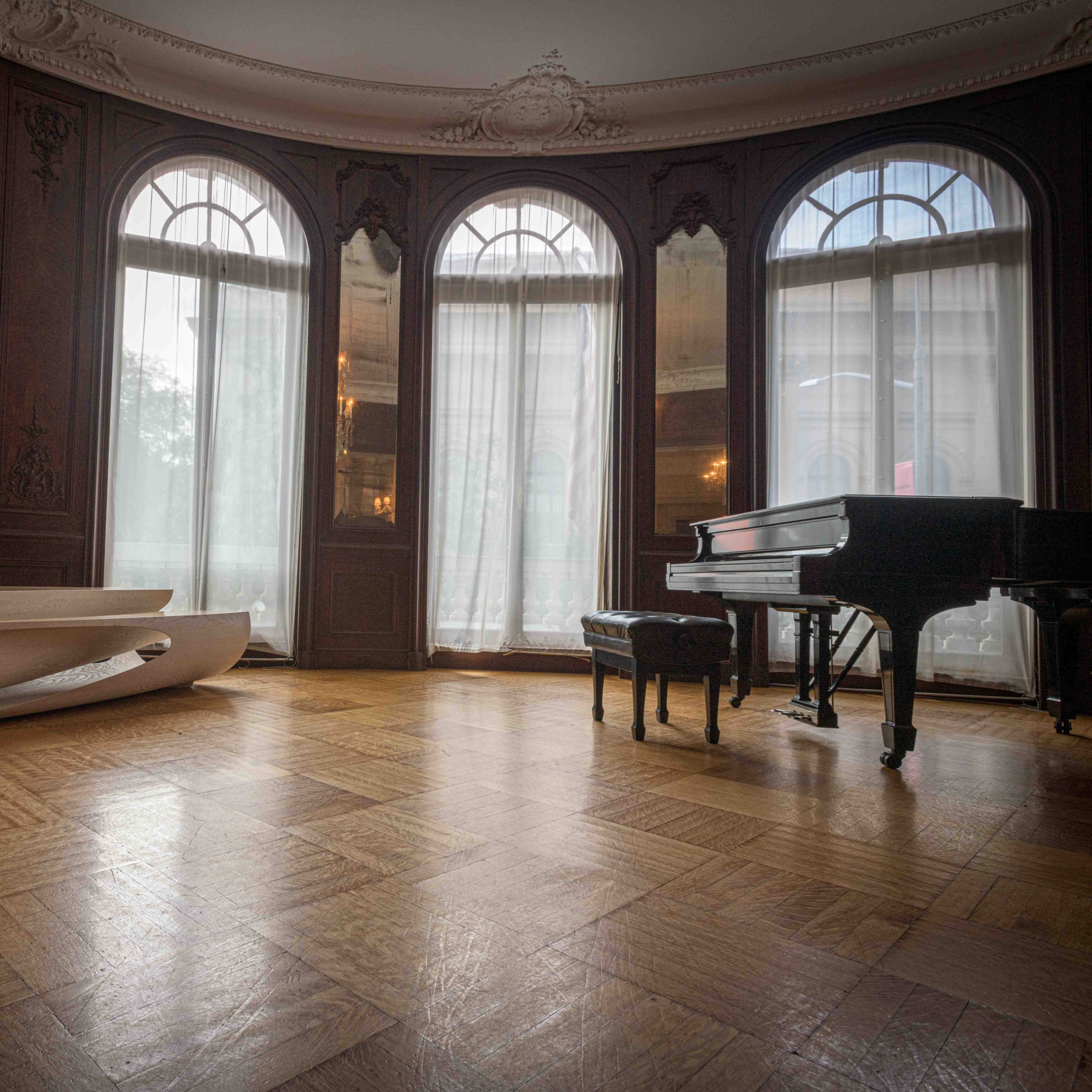 A room with dark wooden walls, large arched windows, a grand piano, and a curved wooden bench on a patterned wooden floor.