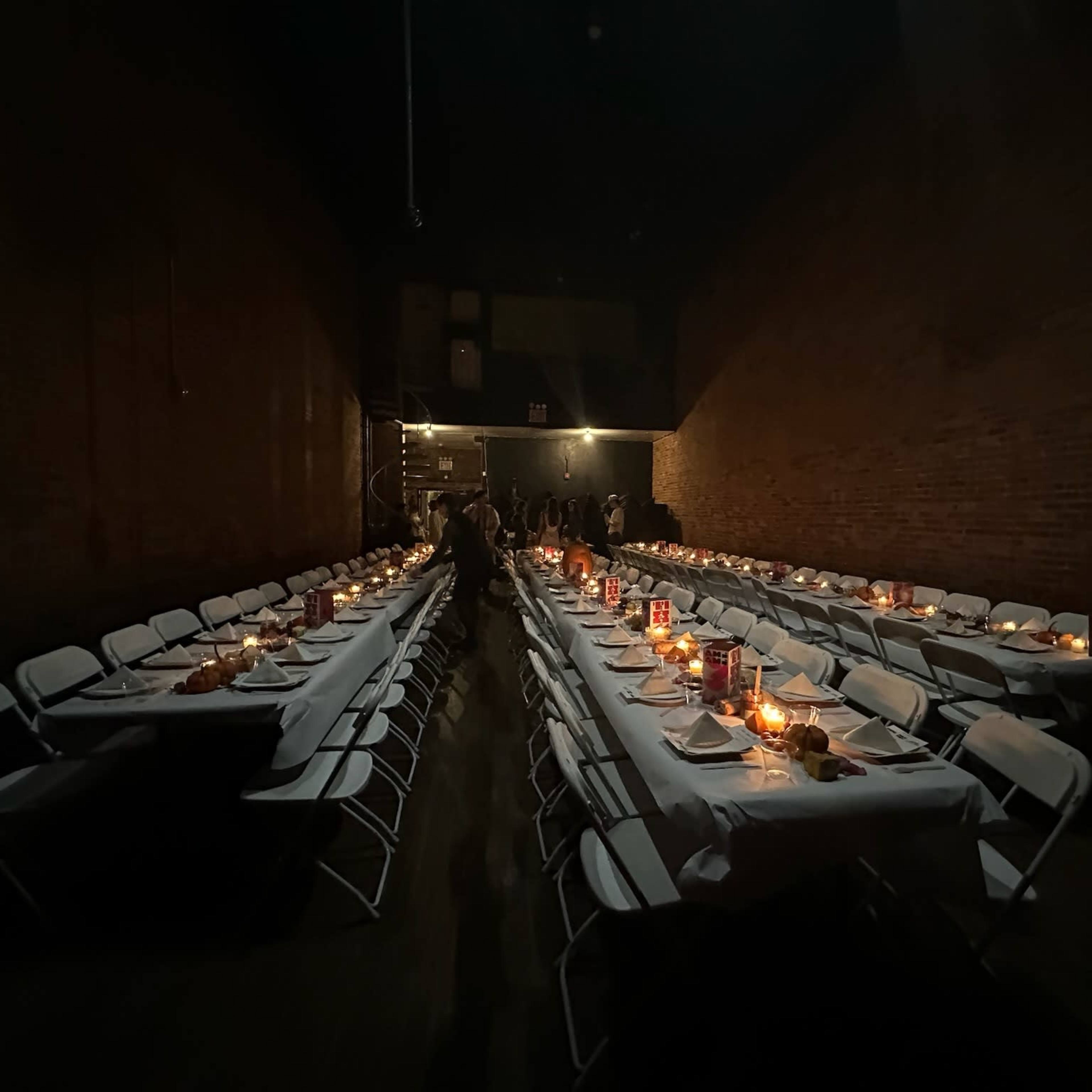A long table with white linens and chairs is arranged in a dimly lit space, with candles and dinnerware set for a meal.