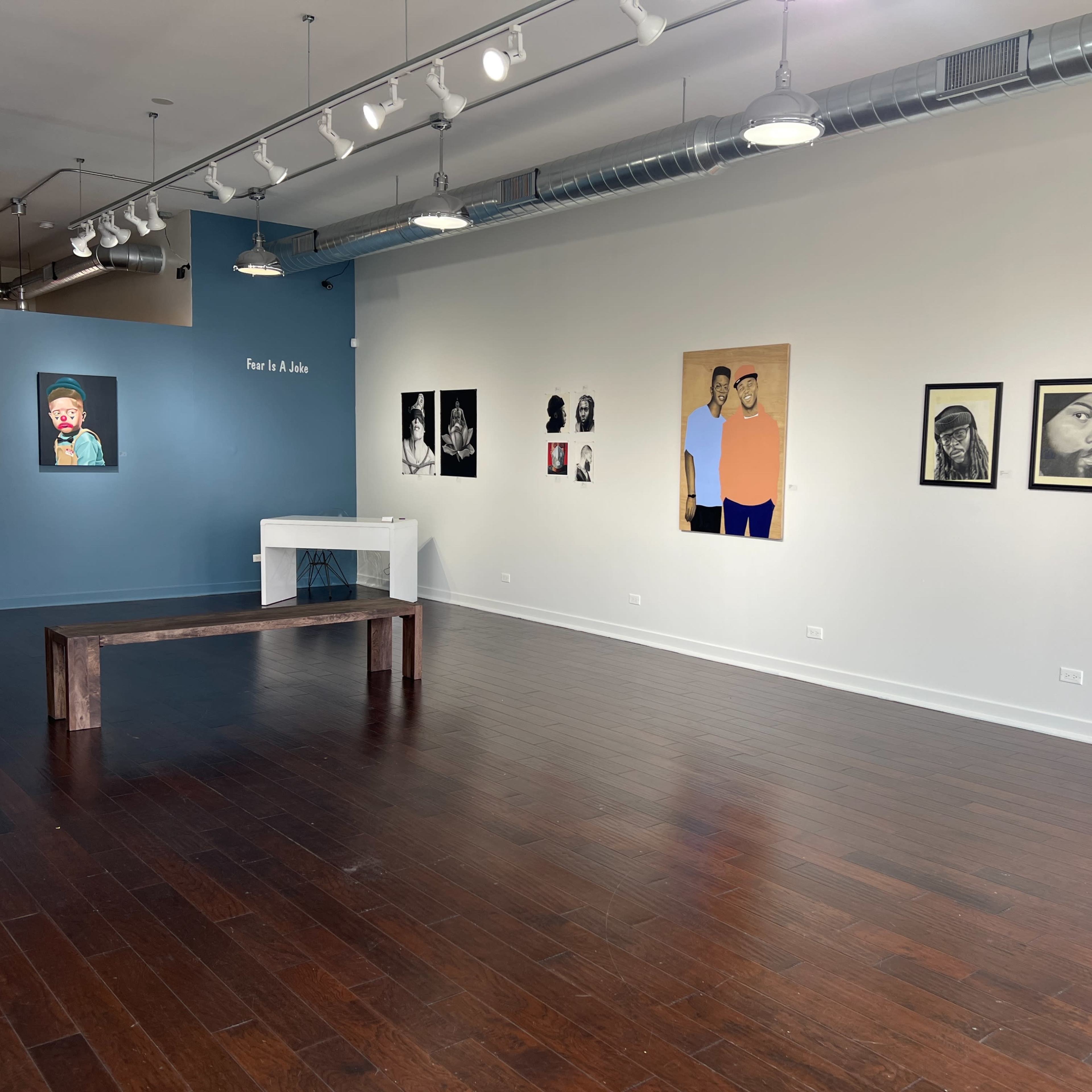 An art gallery with a wooden bench in the center, featuring a blue wall with text and several framed artworks displayed on the white walls.
