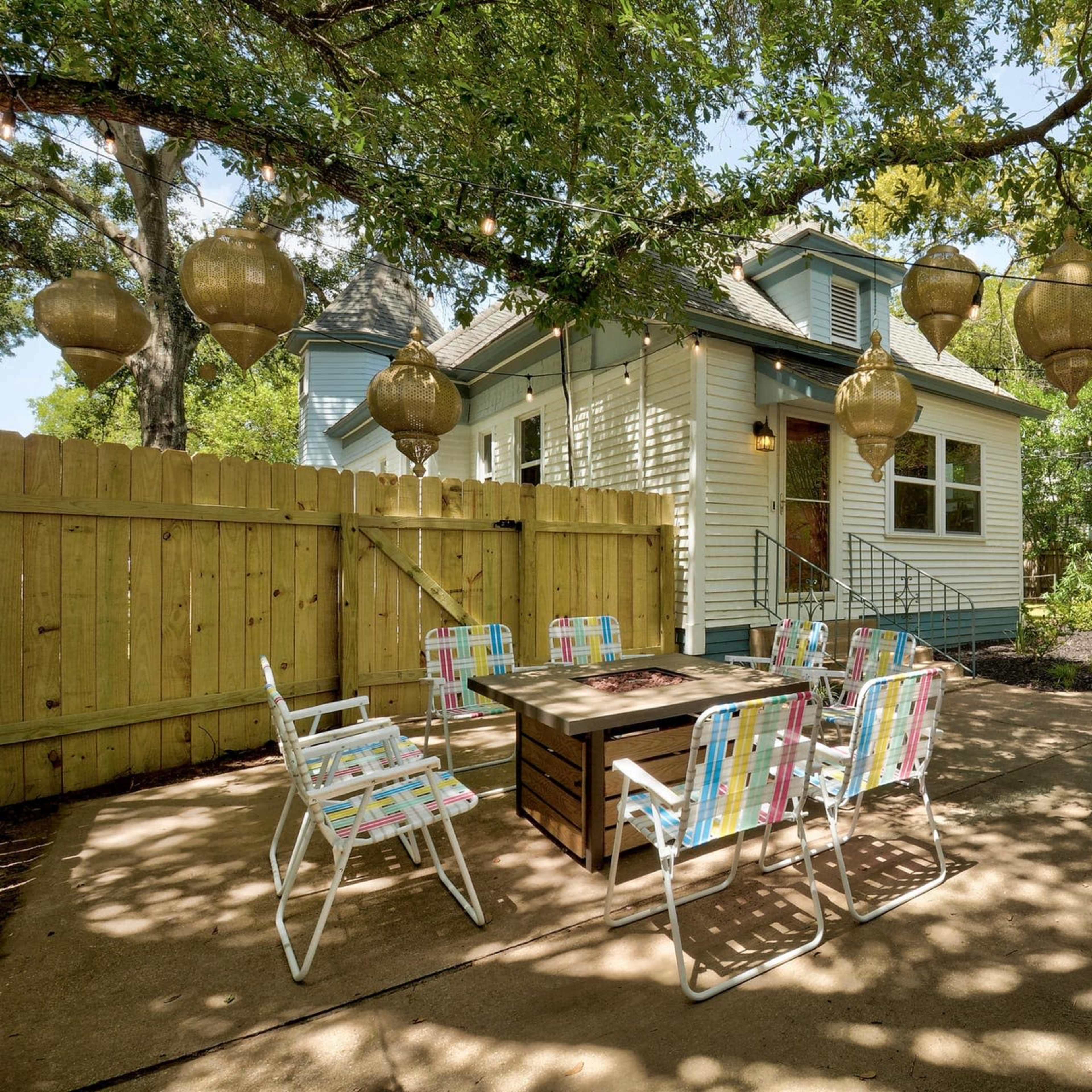 A backyard patio with a table and chairs surrounded by a wooden fence, leading to a house and a gazebo in the distance.
