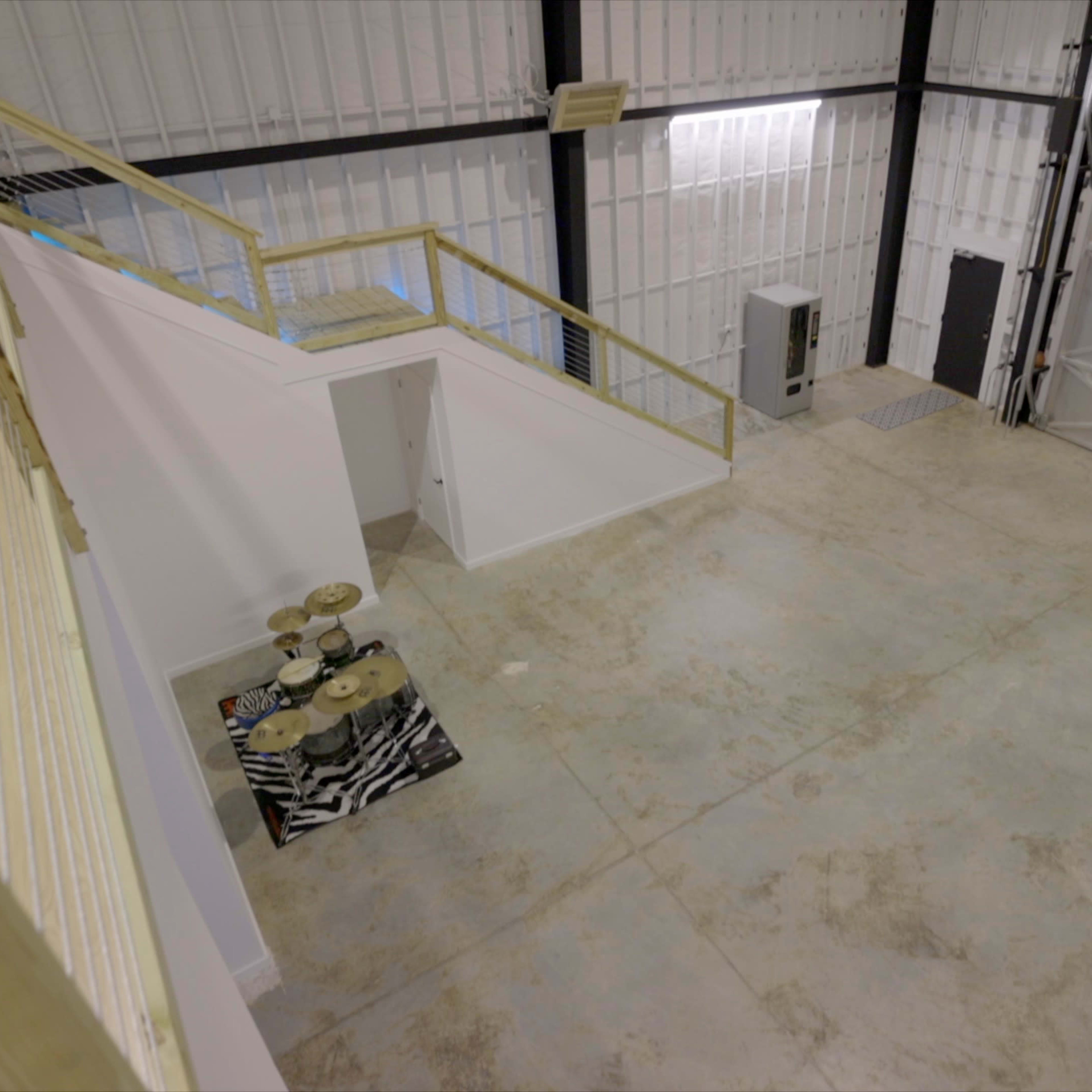 An expansive industrial space features a concrete floor, a staircase leading to a raised platform, and a few items including a drum set on a patterned mat.