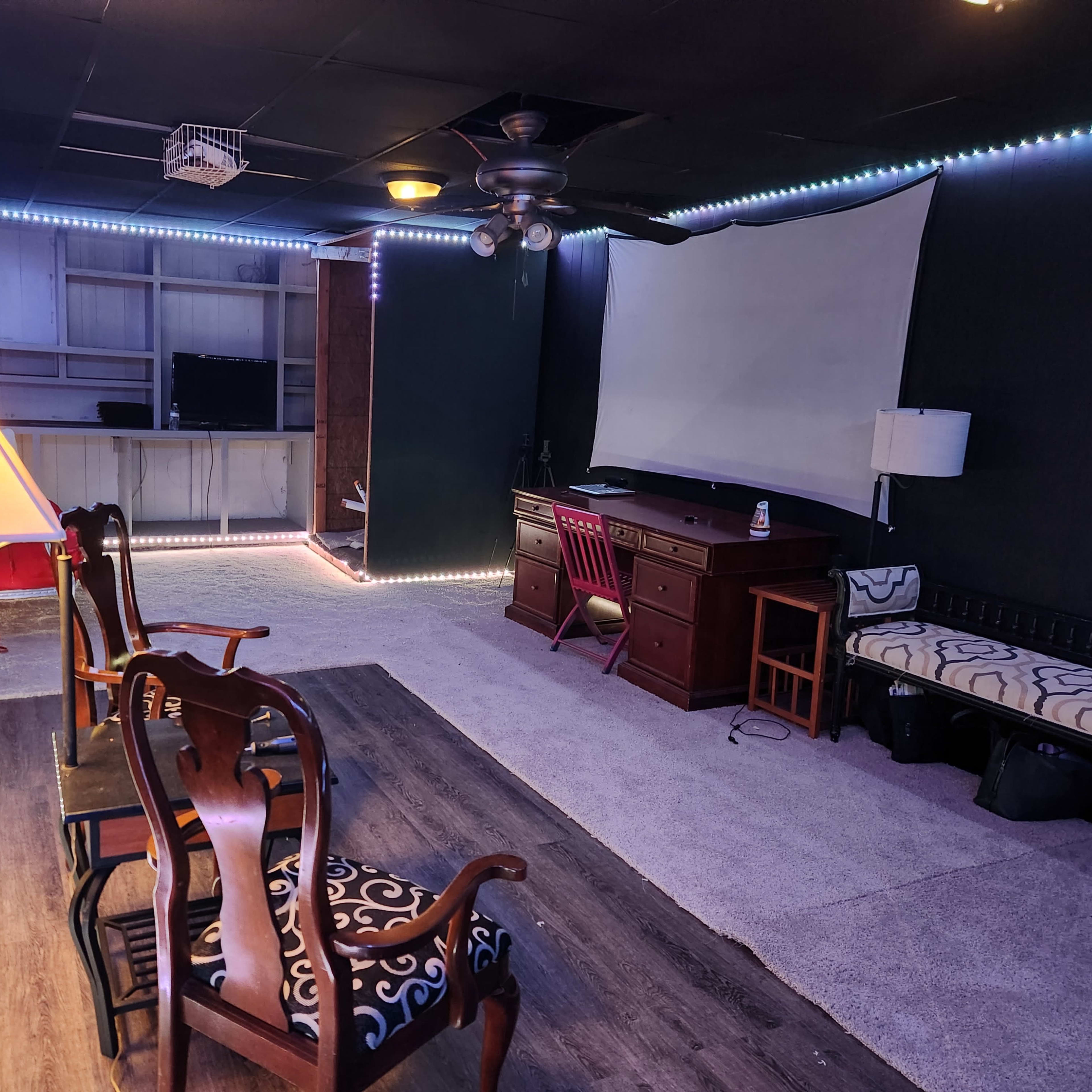 A basement room furnished with chairs, a desk, and a wall-mounted screen, illuminated by LED lights along the perimeter.