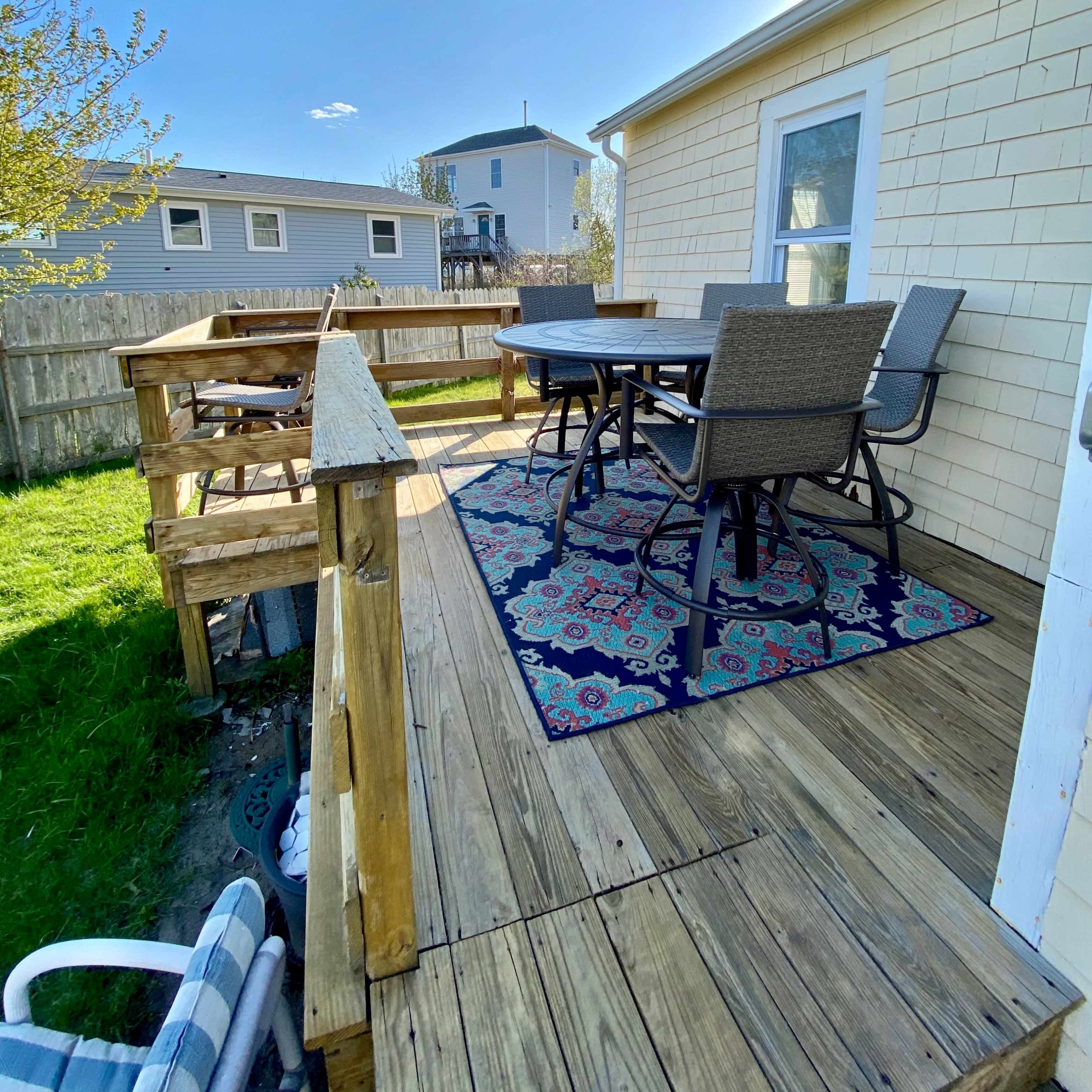 A wooden deck with a patterned rug and a dining set, overlooking a grassy yard and neighboring houses.