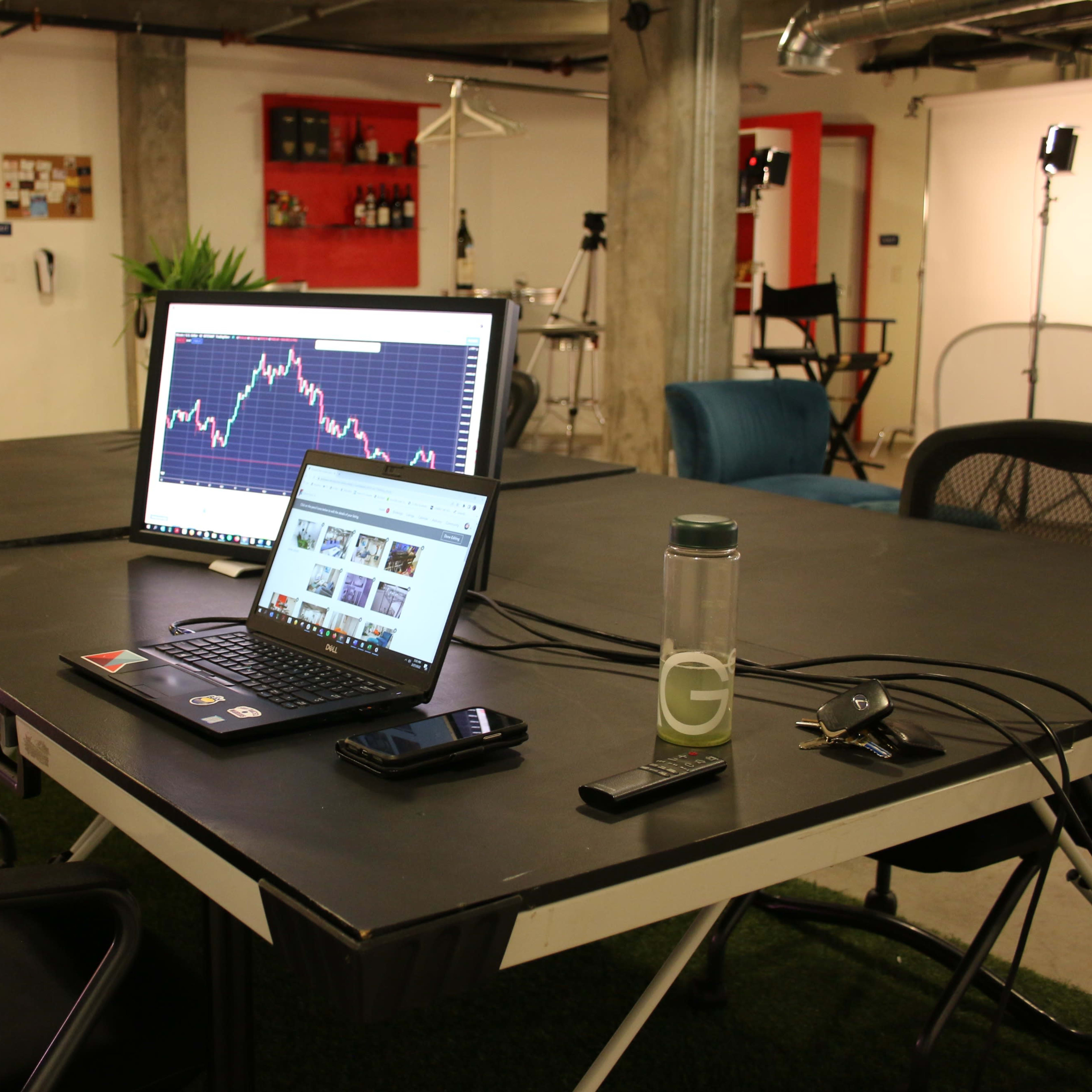 A workspace contains a laptop and a monitor displaying charts, surrounded by chairs and potted plants.