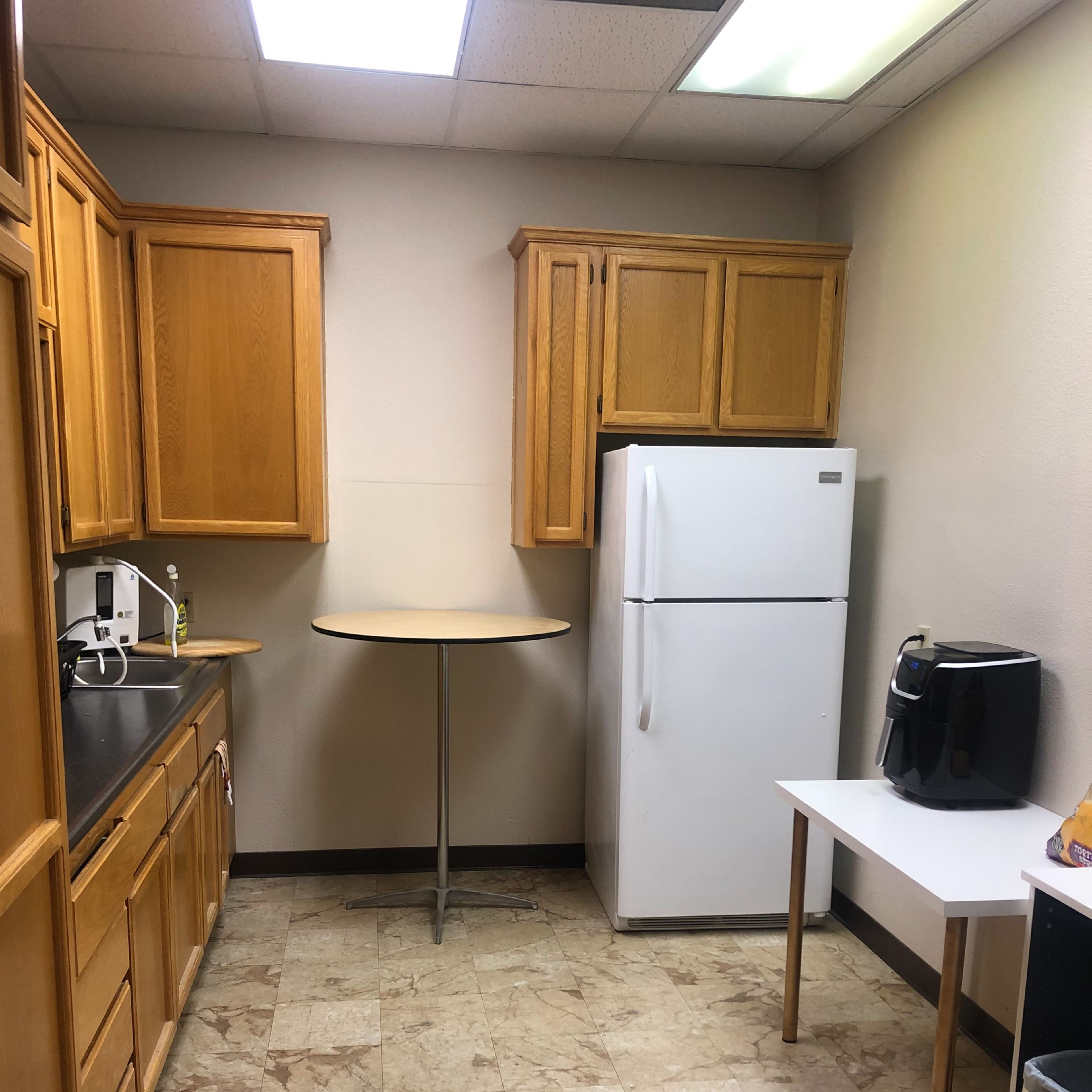 A small kitchen with wooden cabinets, a white refrigerator, a circular table, and a coffee maker on a white table.