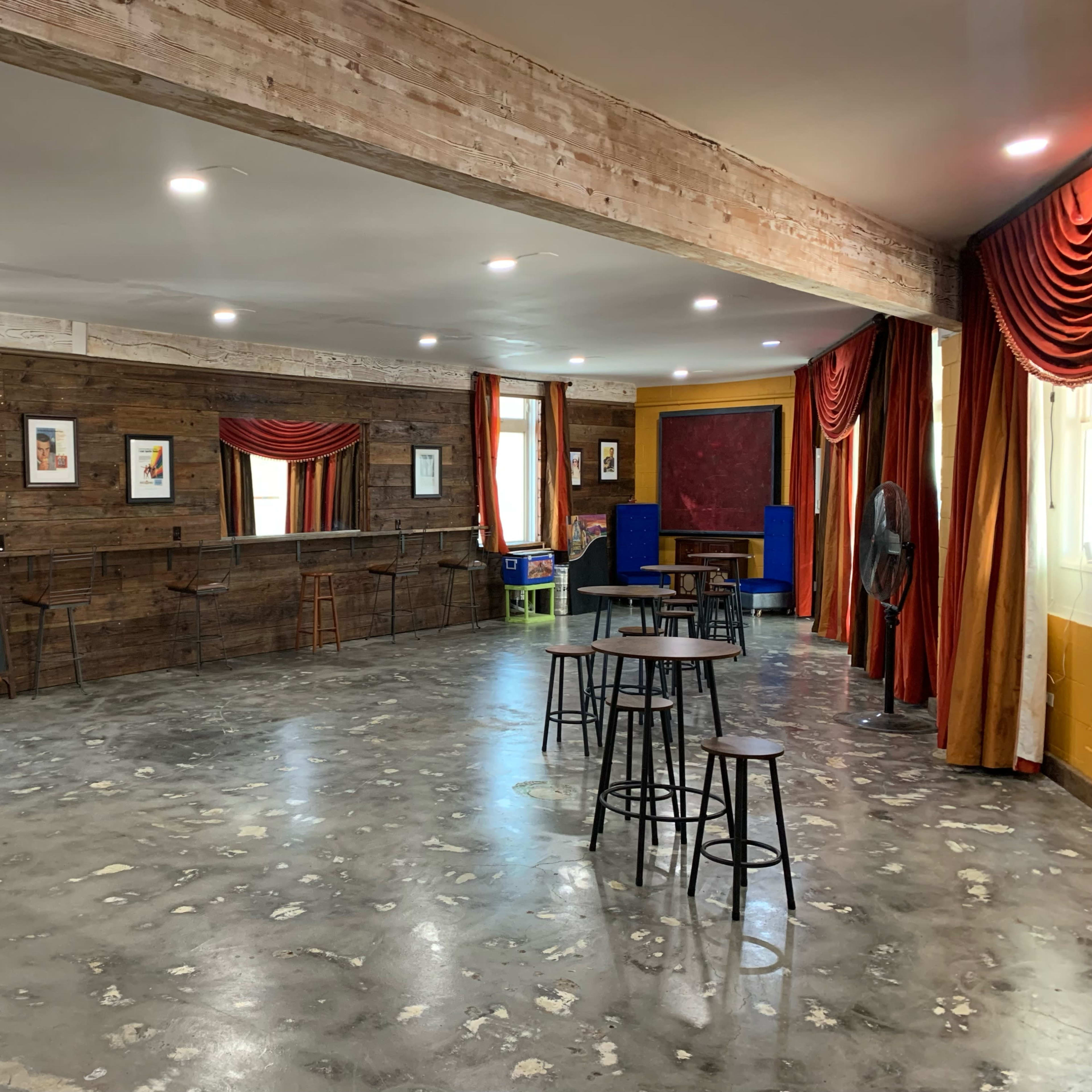 A spacious room with wooden walls, several tables and chairs, and large windows draped with curtains.