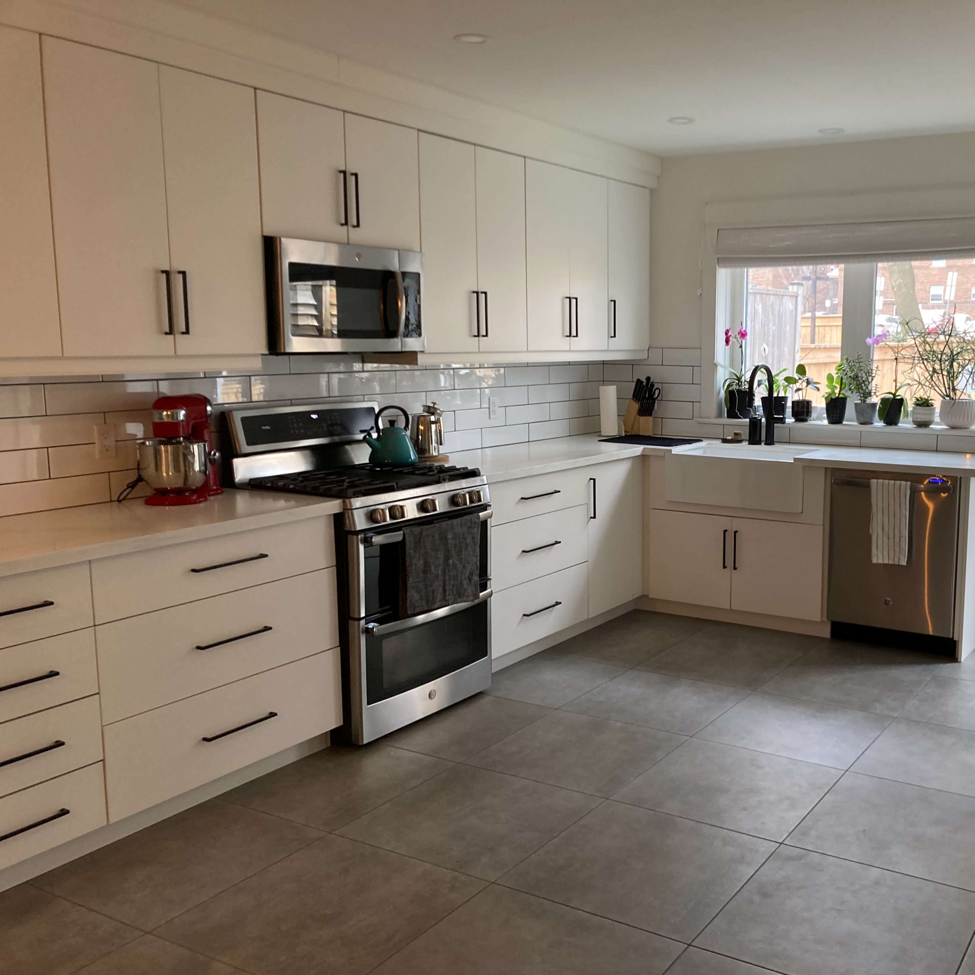 A modern kitchen features white cabinets, stainless steel appliances, and a large window with plants on the sill.