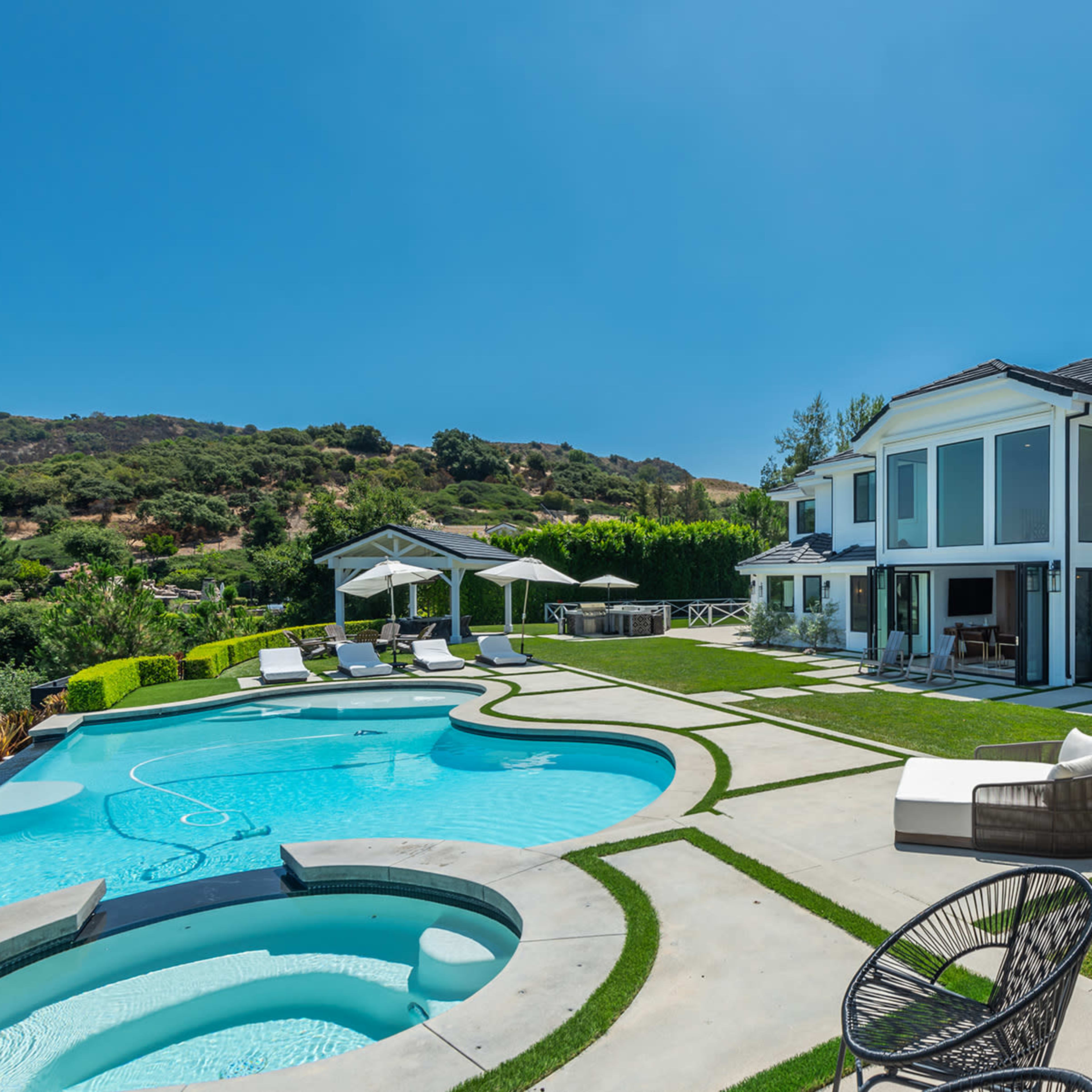 A modern house with large windows overlooks a swimming pool and landscaped yard in a sunny setting.
