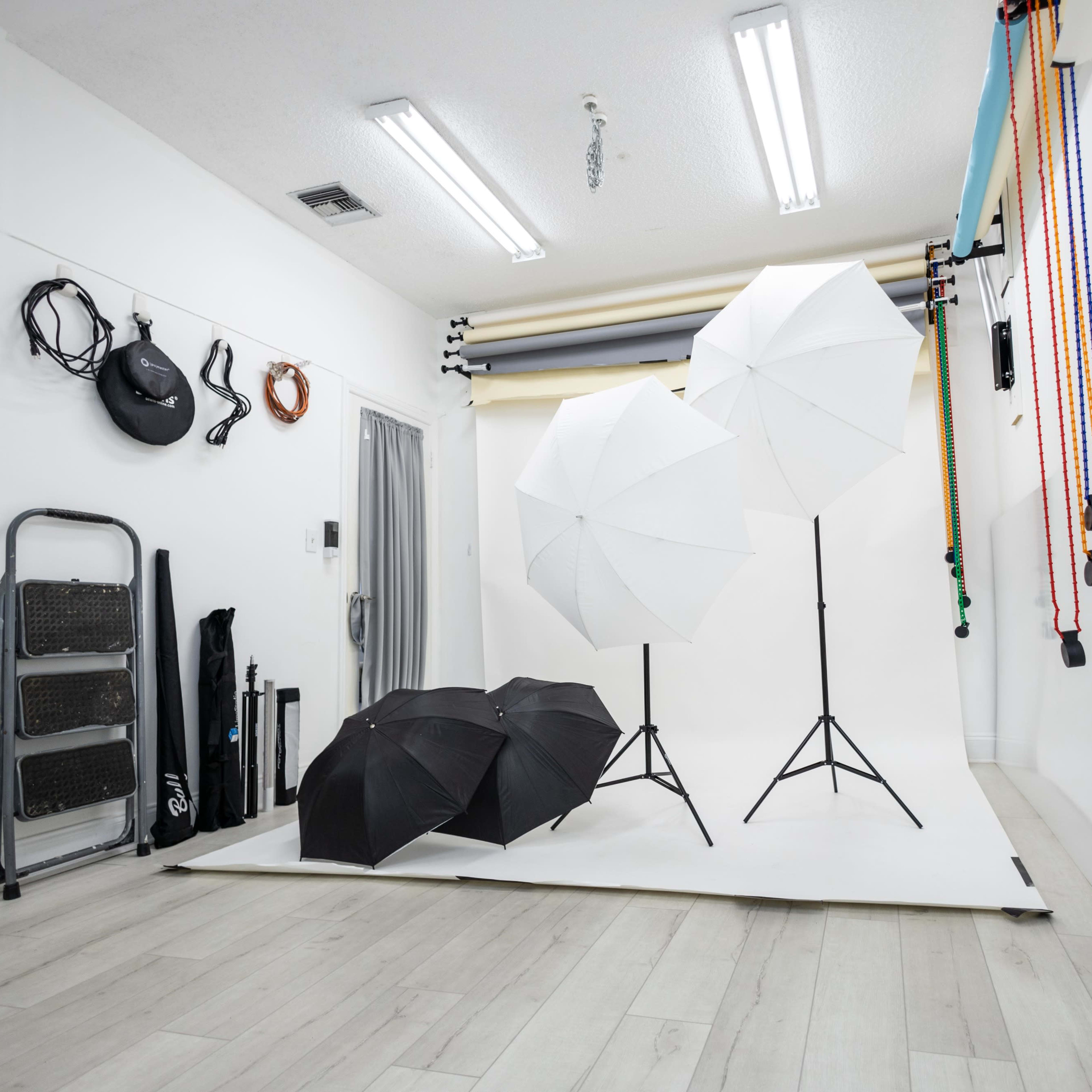 A photography studio with two white umbrellas and one black umbrella on tripods, situated against a plain backdrop with various props and equipment along the walls.