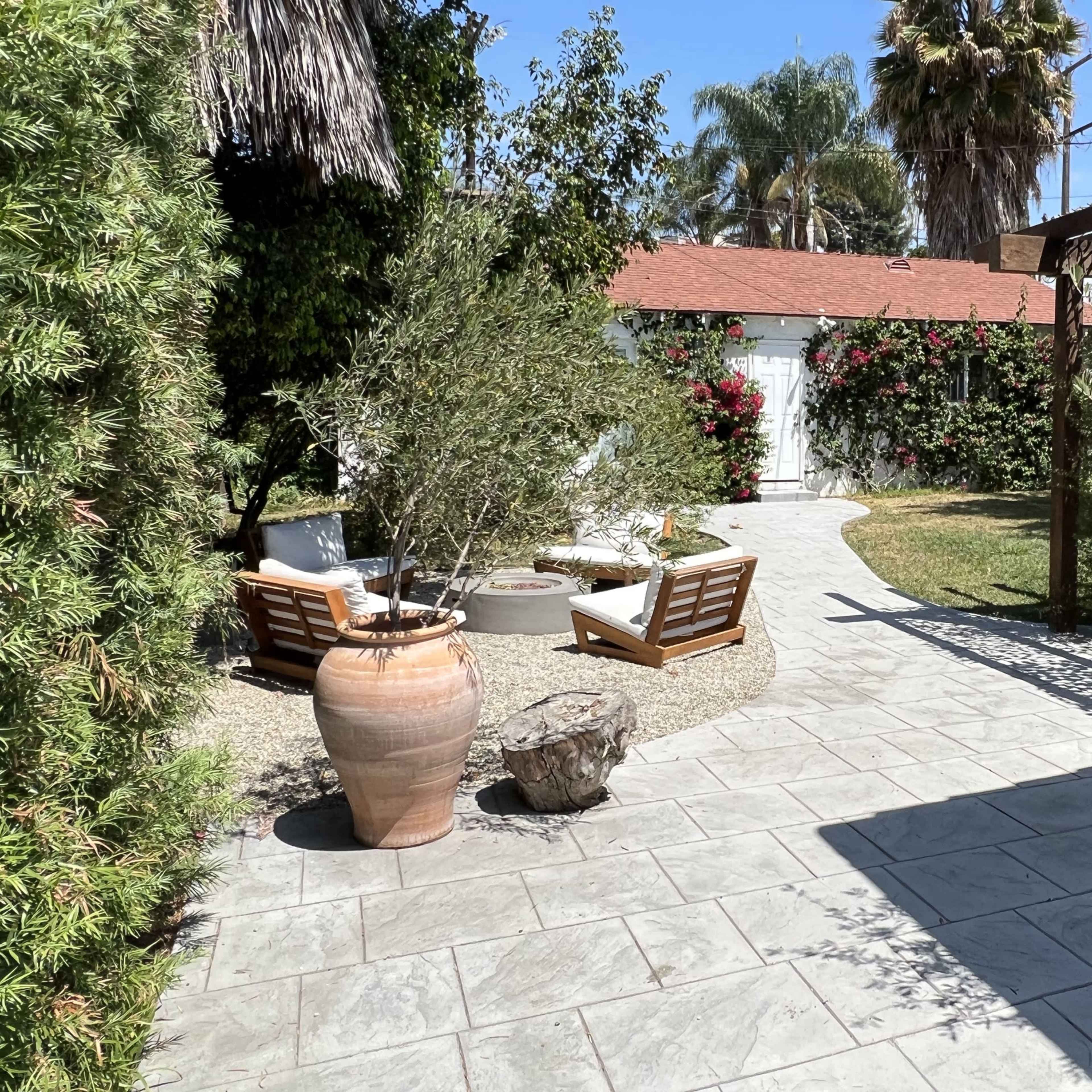 A stone pathway leads through a garden with potted plants and lounge chairs, surrounded by greenery and flowering bushes.