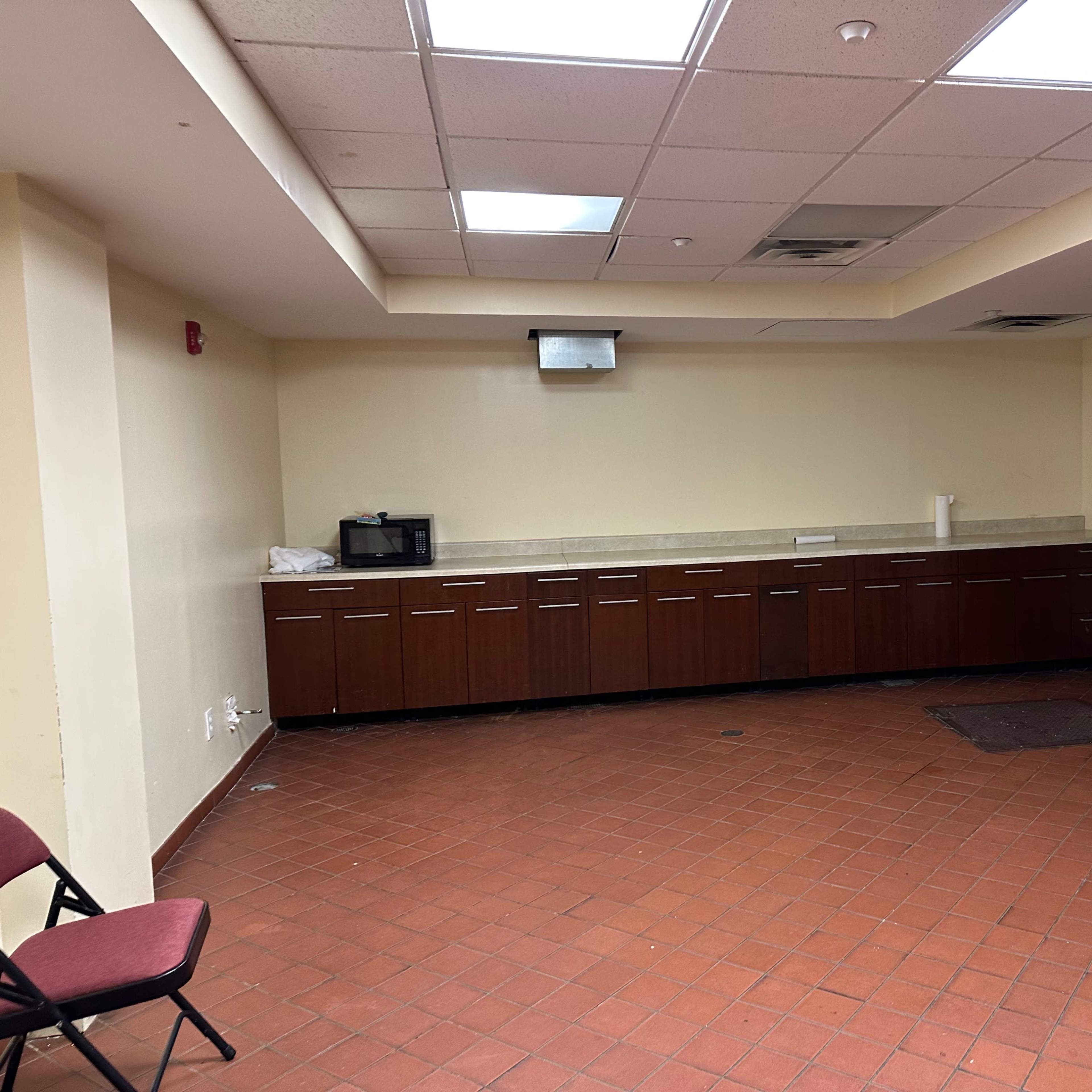 A kitchen area with cabinets, a countertop, a microwave, and a refrigerator on one side, with a few empty chairs and a blue tarp on the floor.