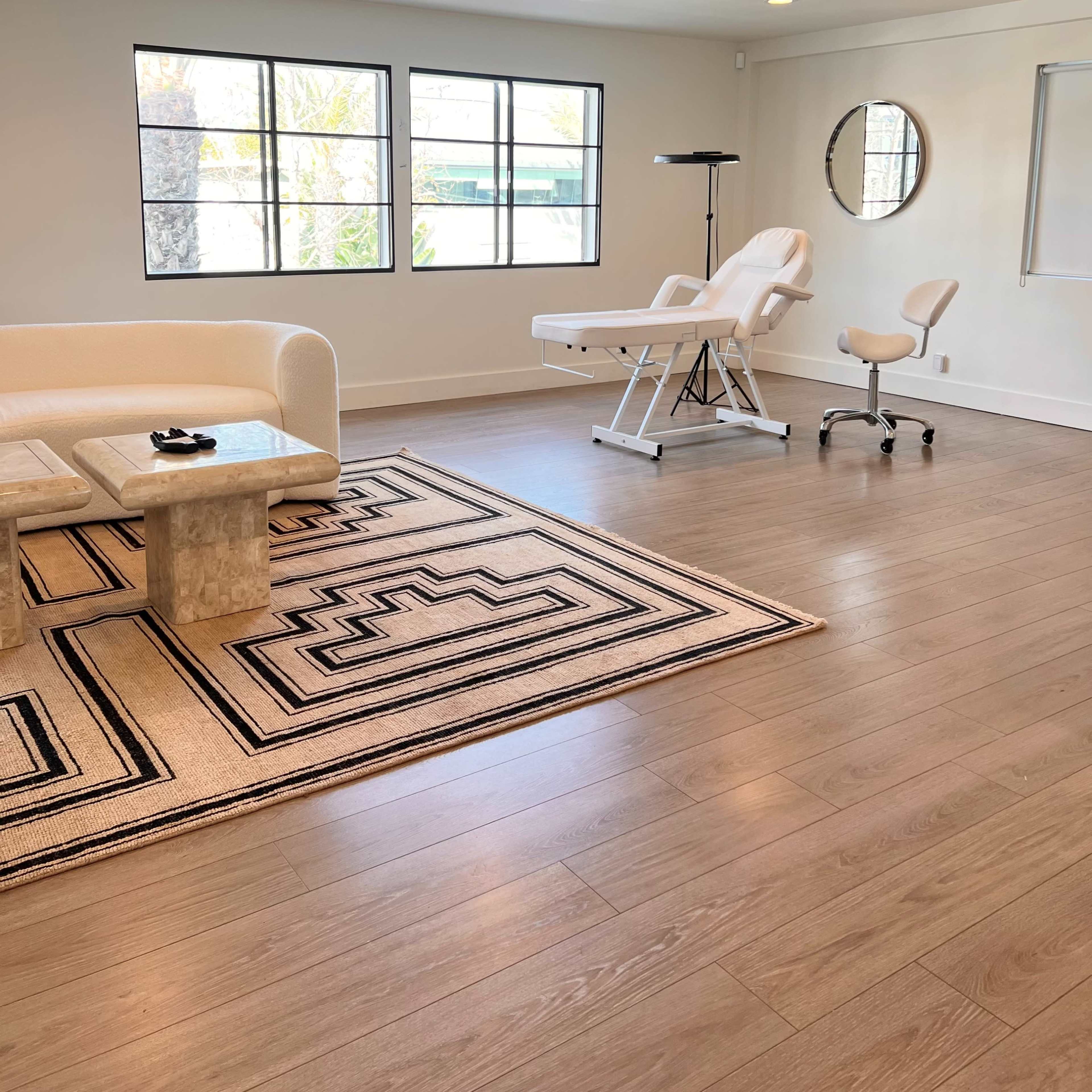 A bright, minimalist treatment room featuring a white couch, a marble coffee table, and a treatment chair beside a large window.
