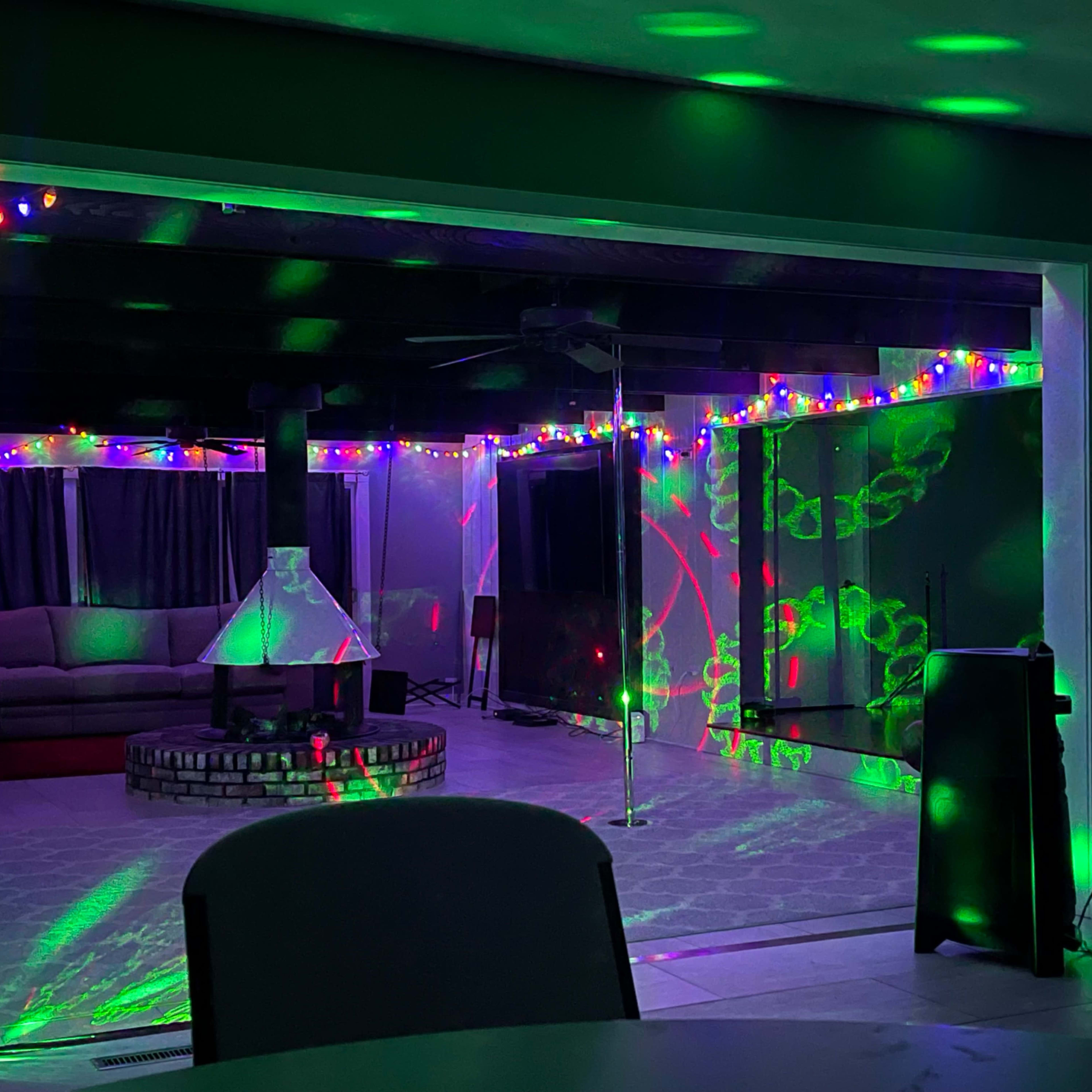 A spacious indoor area is decorated with colorful lights, featuring a seating arrangement, a central firepit, and a dance pole.