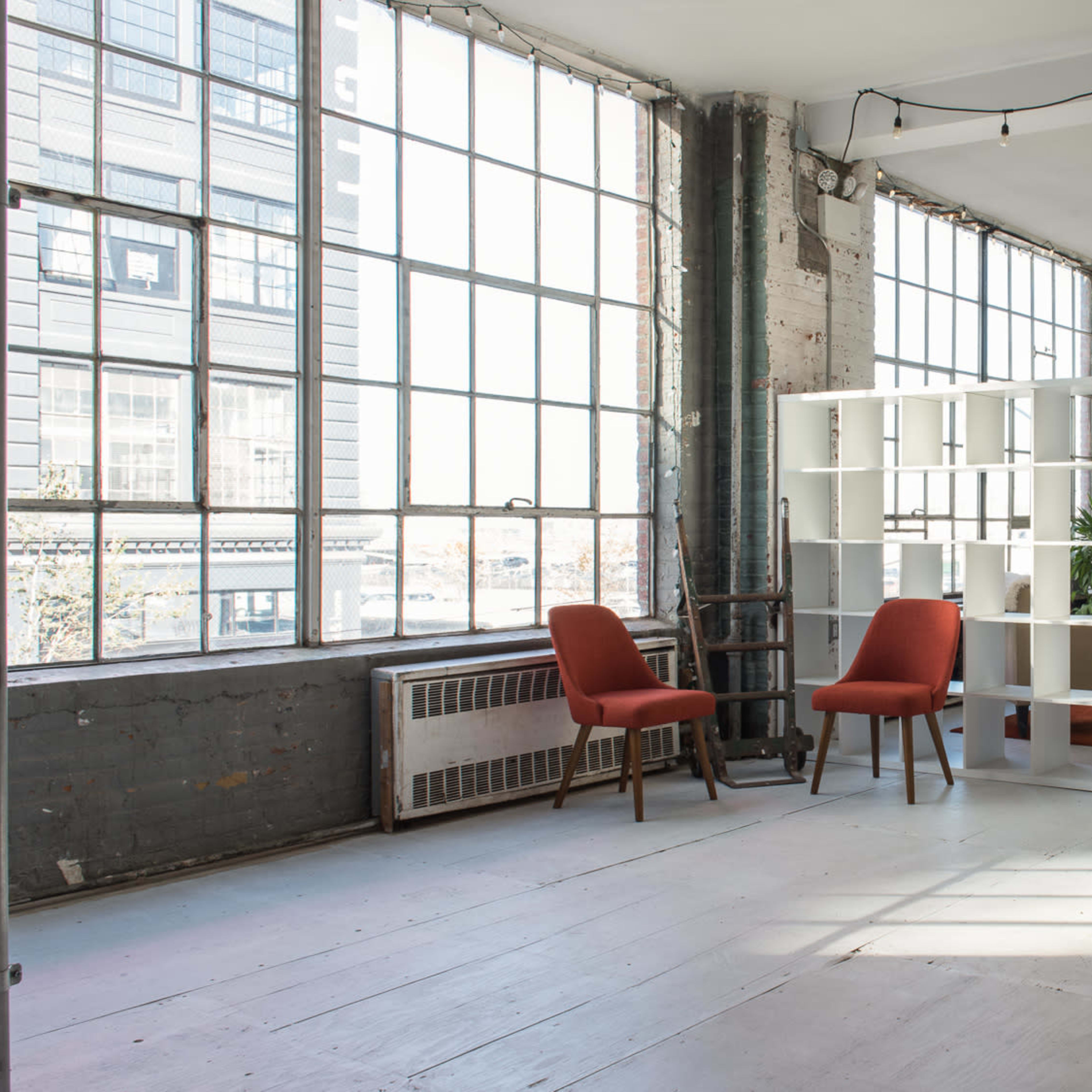 A spacious room features large windows, a bookshelf, two red chairs, and a couch on a rug.