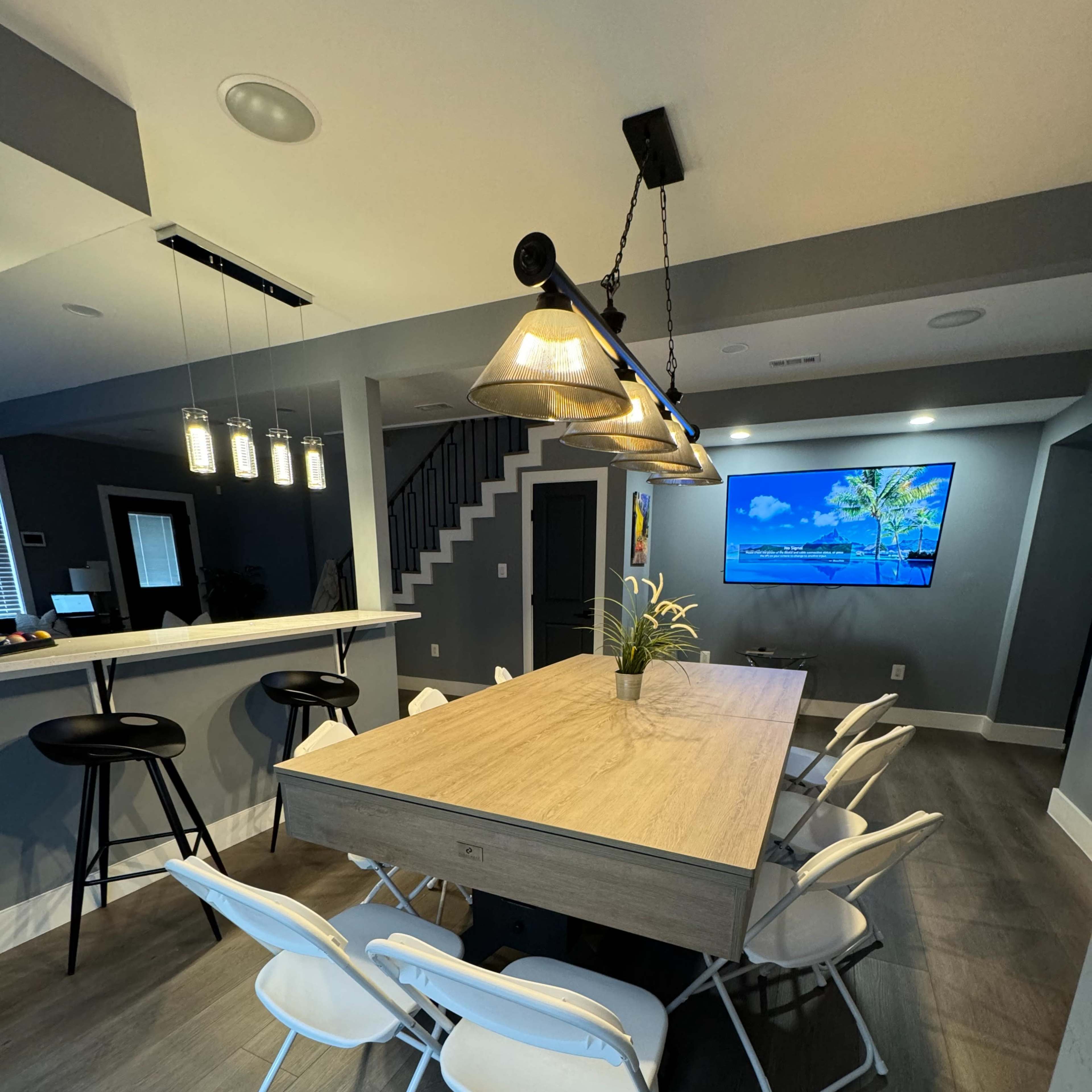 A modern dining area featuring a large wooden table surrounded by white folding chairs, with a bar counter in the background and a wall-mounted television.