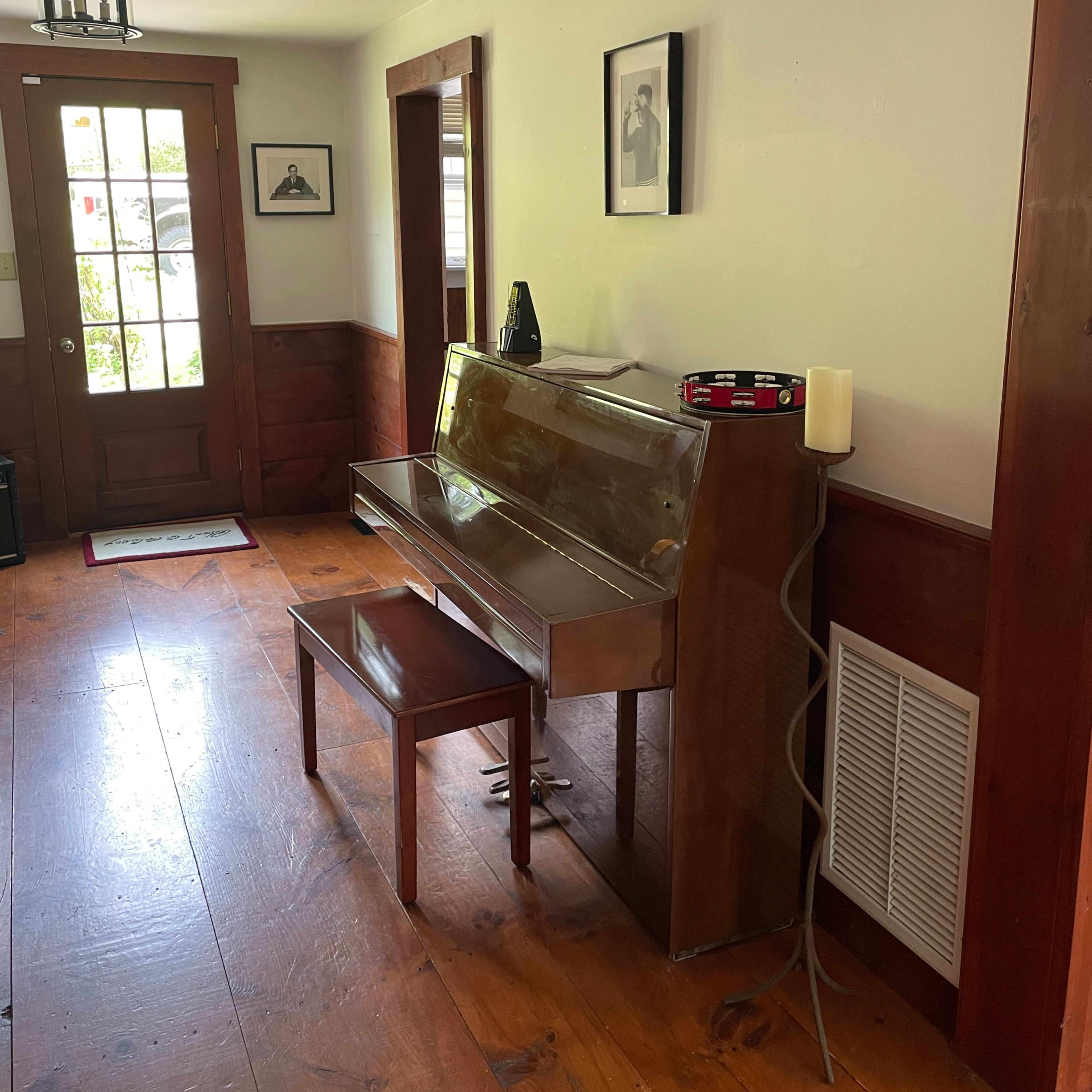 A wooden piano with a matching bench positioned in a well-lit entryway, beside a door and framed photographs on the wall.