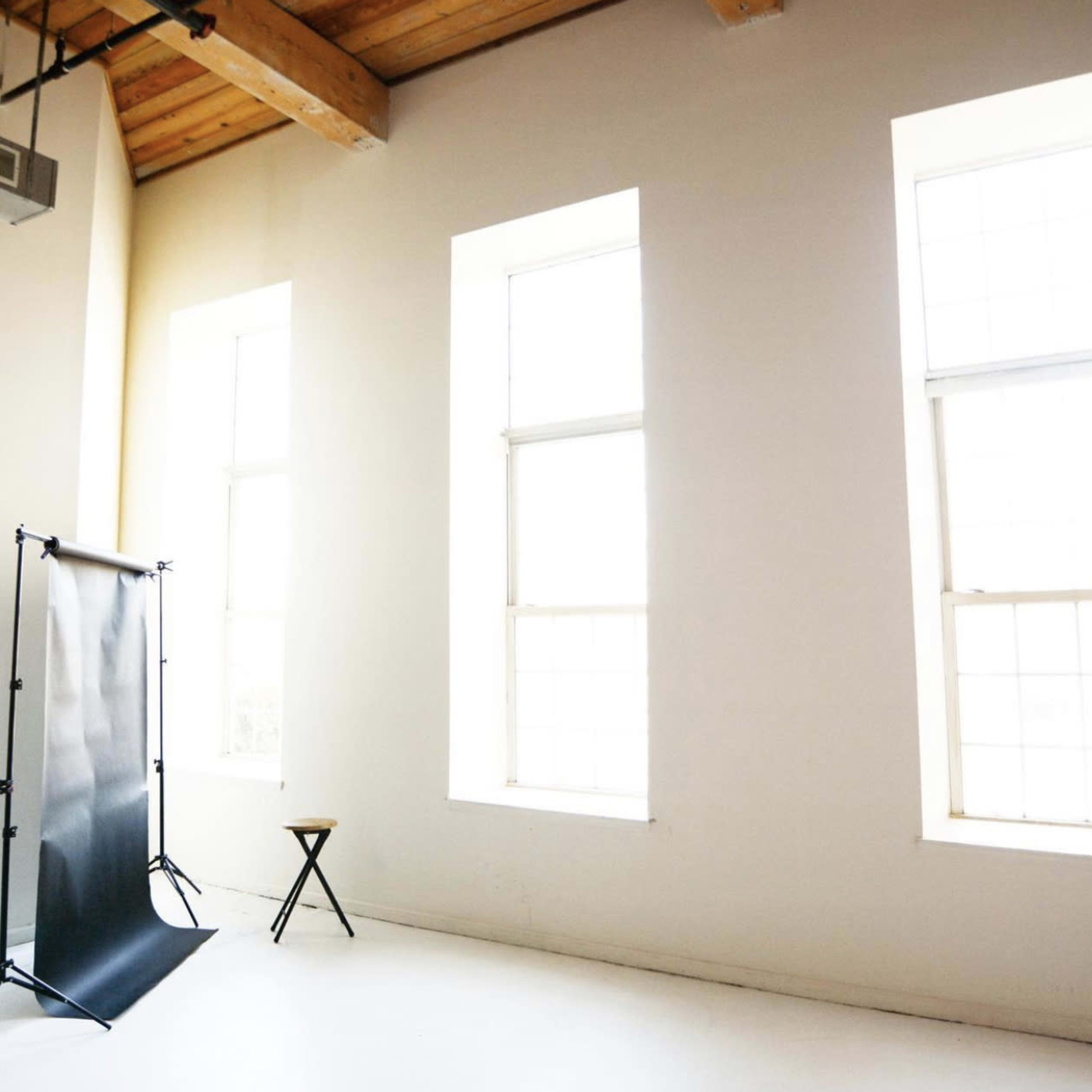 A well-lit room with three large windows, a black backdrop on a stand, and a wooden stool.
