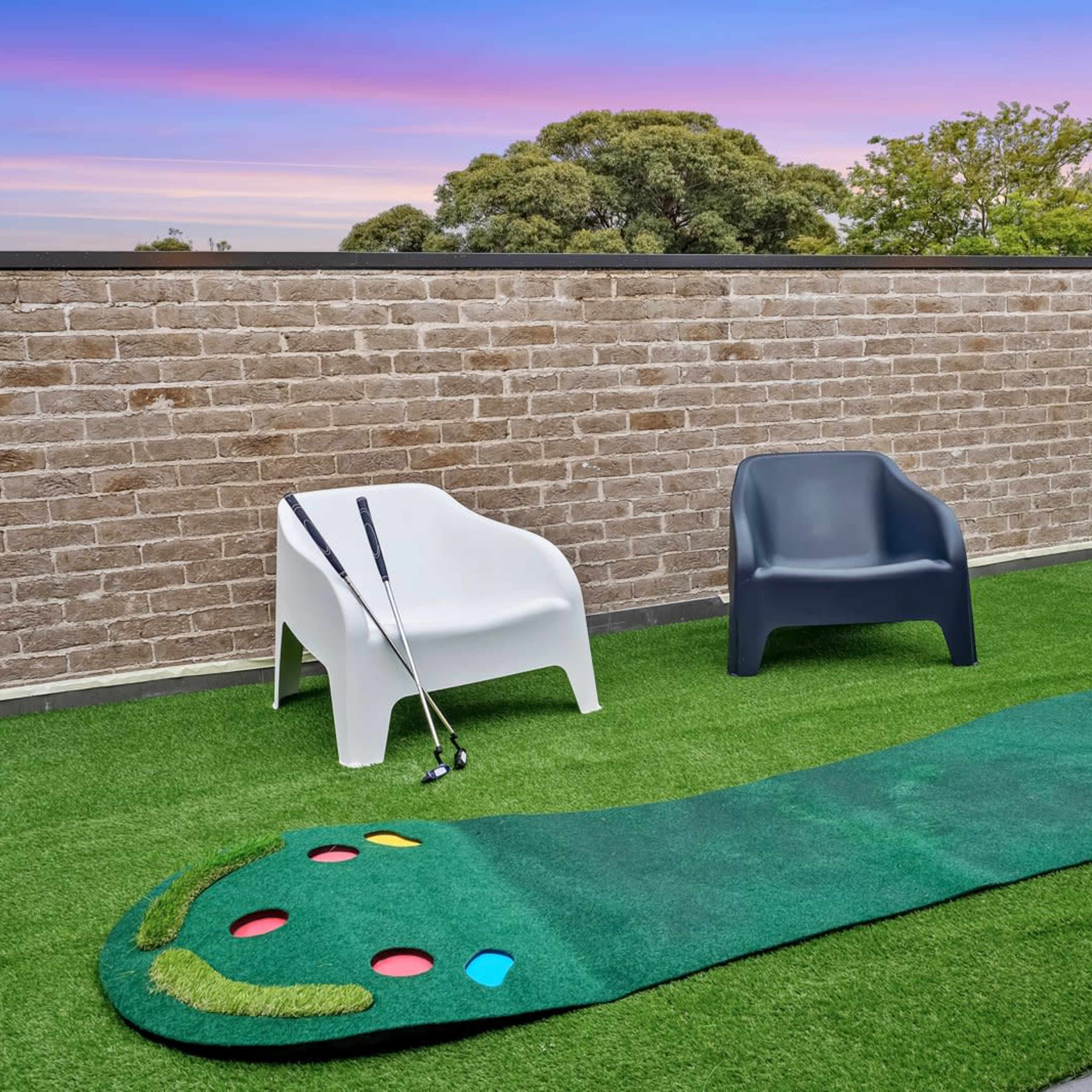 A putting green with colorful holes, two modern chairs, and golf clubs resting against a brick wall under a colorful sky.