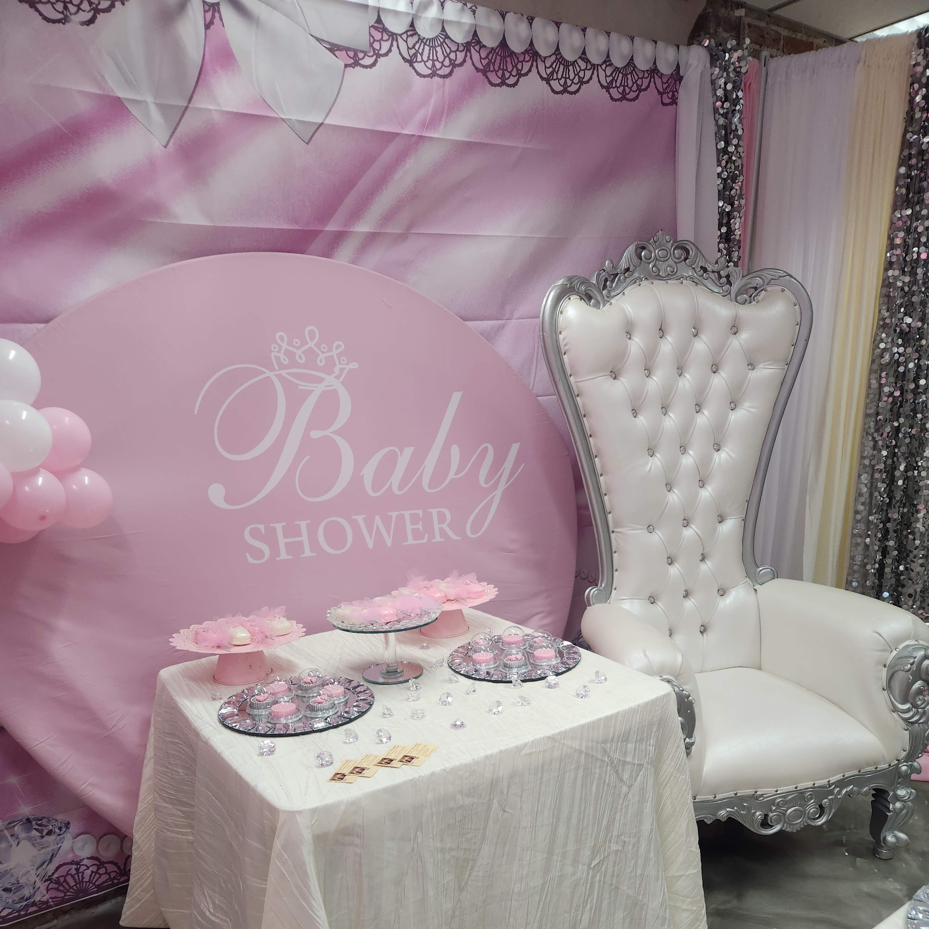 A decorated baby shower setting with a large pink backdrop, a white table covered with a cloth, and an ornate silver throne chair.