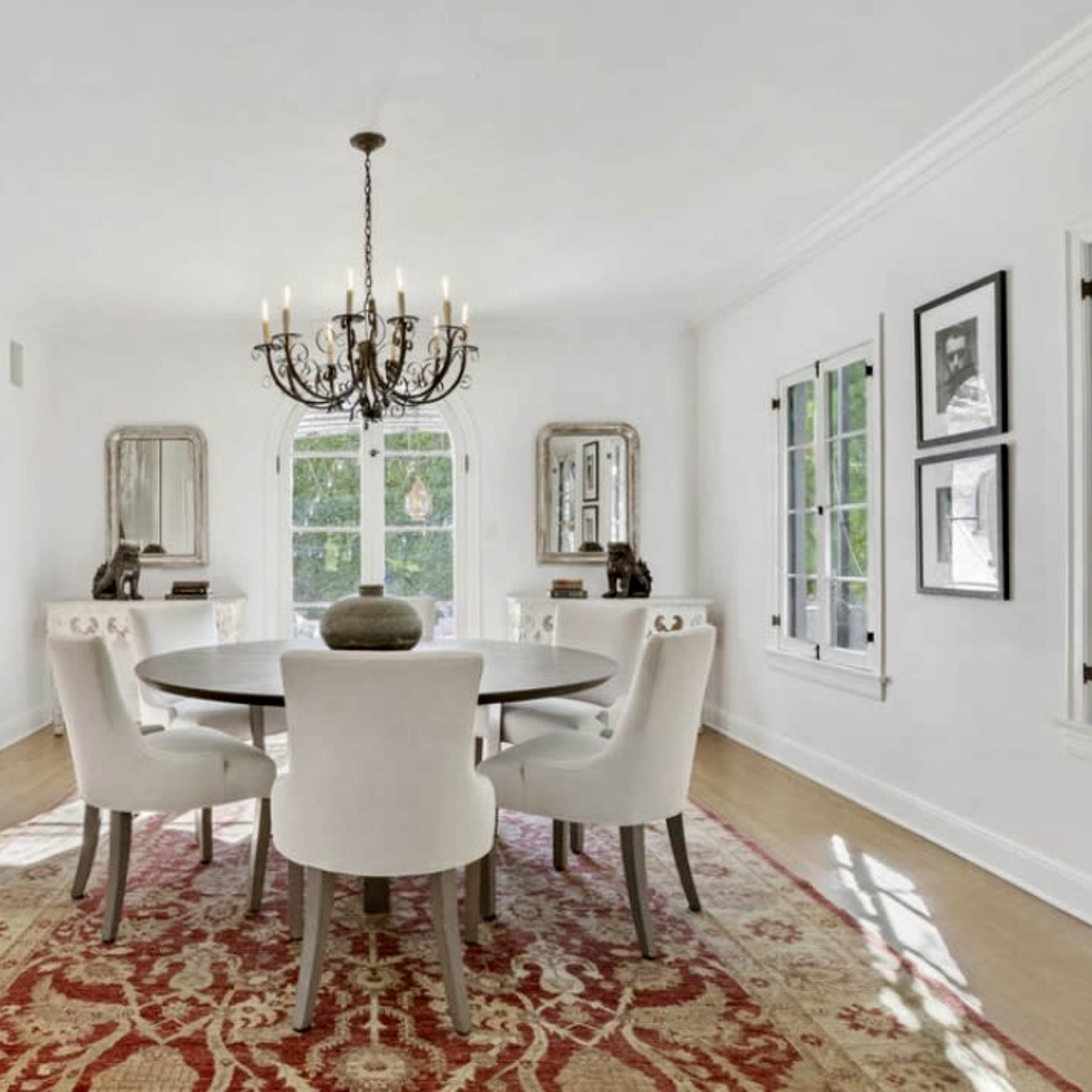 A bright dining room featuring a circular table surrounded by upholstered chairs, a chandelier overhead, and large windows letting in natural light, with a decorative rug on the floor and framed art on the walls.
