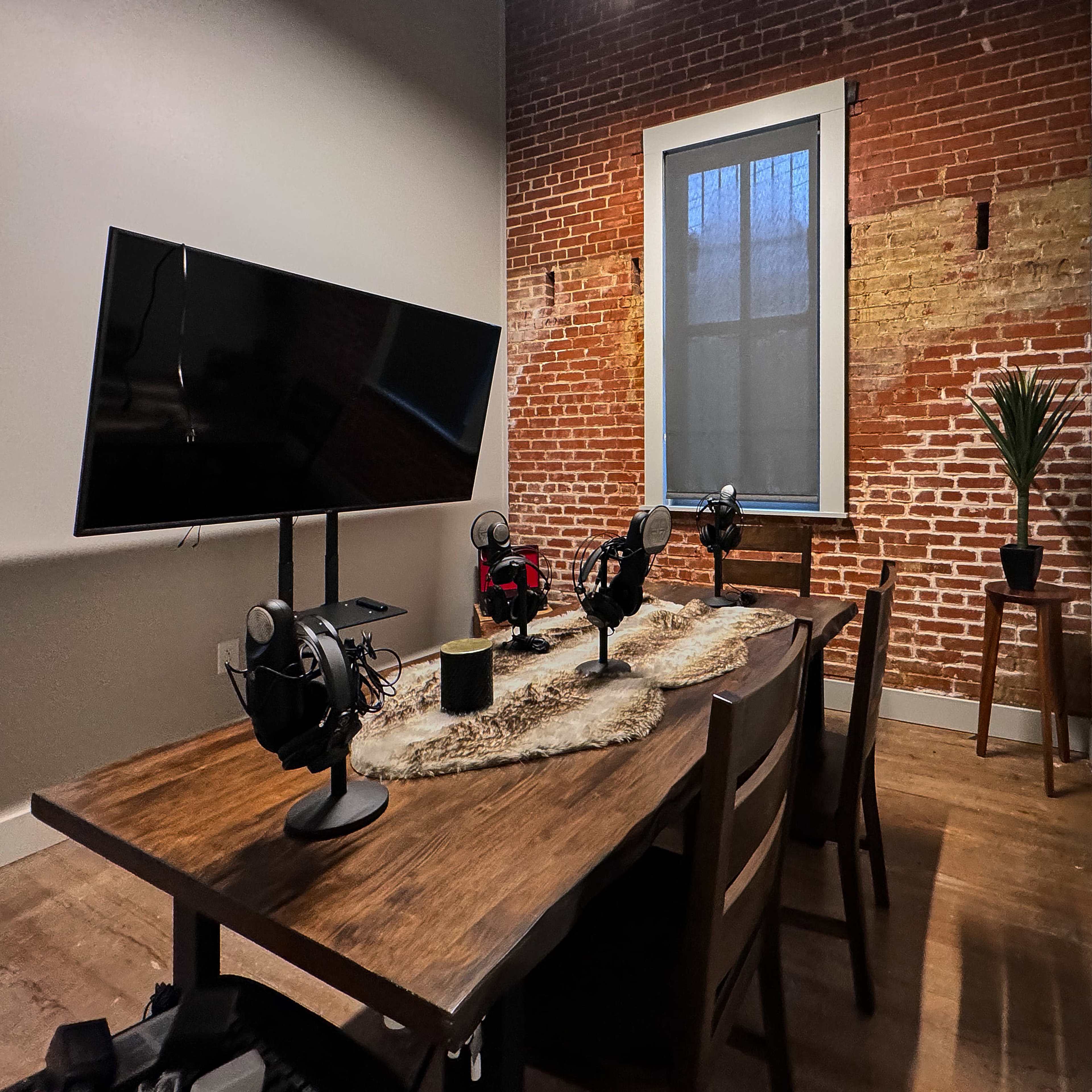 A small meeting room with a wooden table, four microphones on the table, a television mounted on the wall, and a large window with a brick wall backdrop.