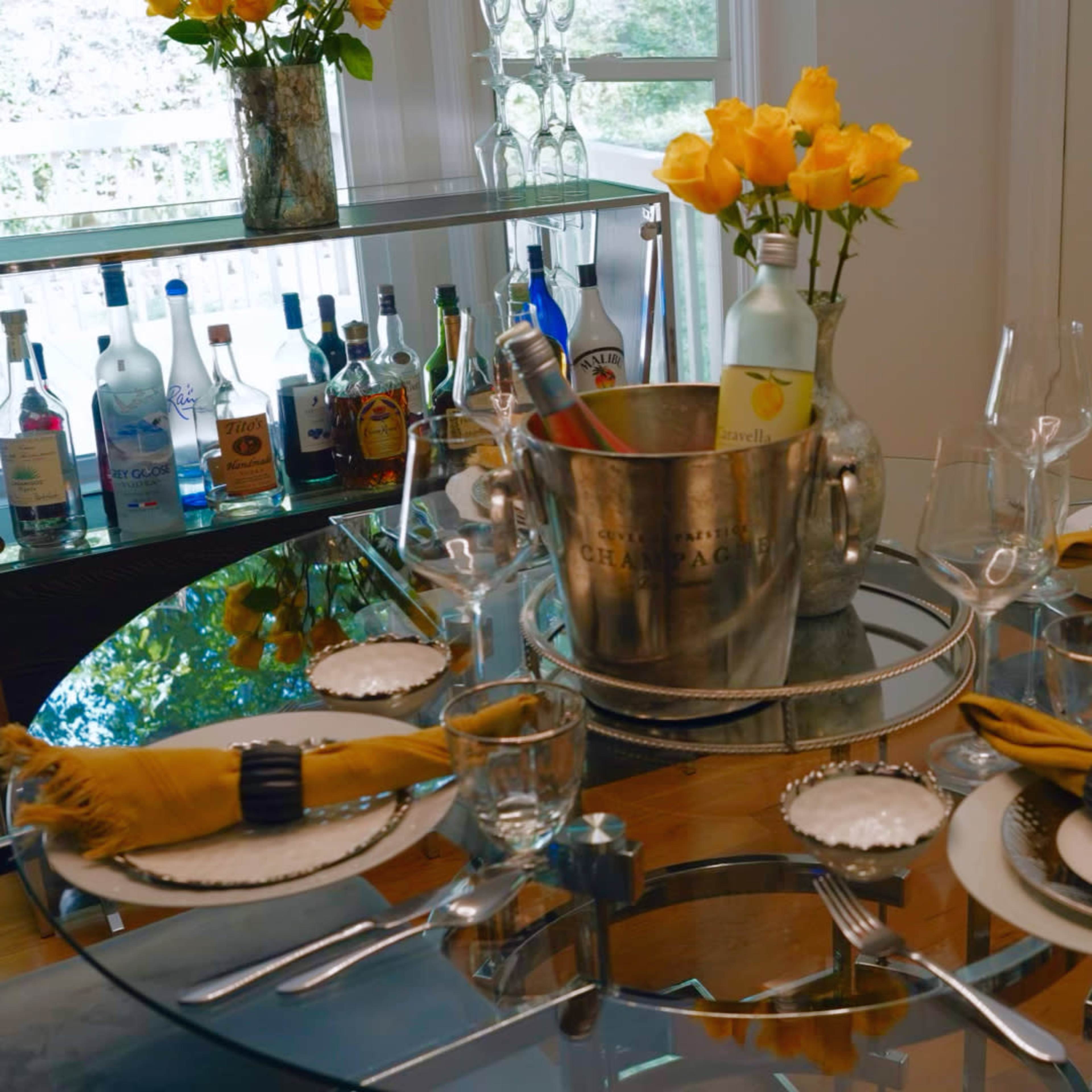A dining table is elegantly set with glassware, napkins, and plates, accompanied by a bucket of drinks and a vase of yellow roses in a well-lit room.