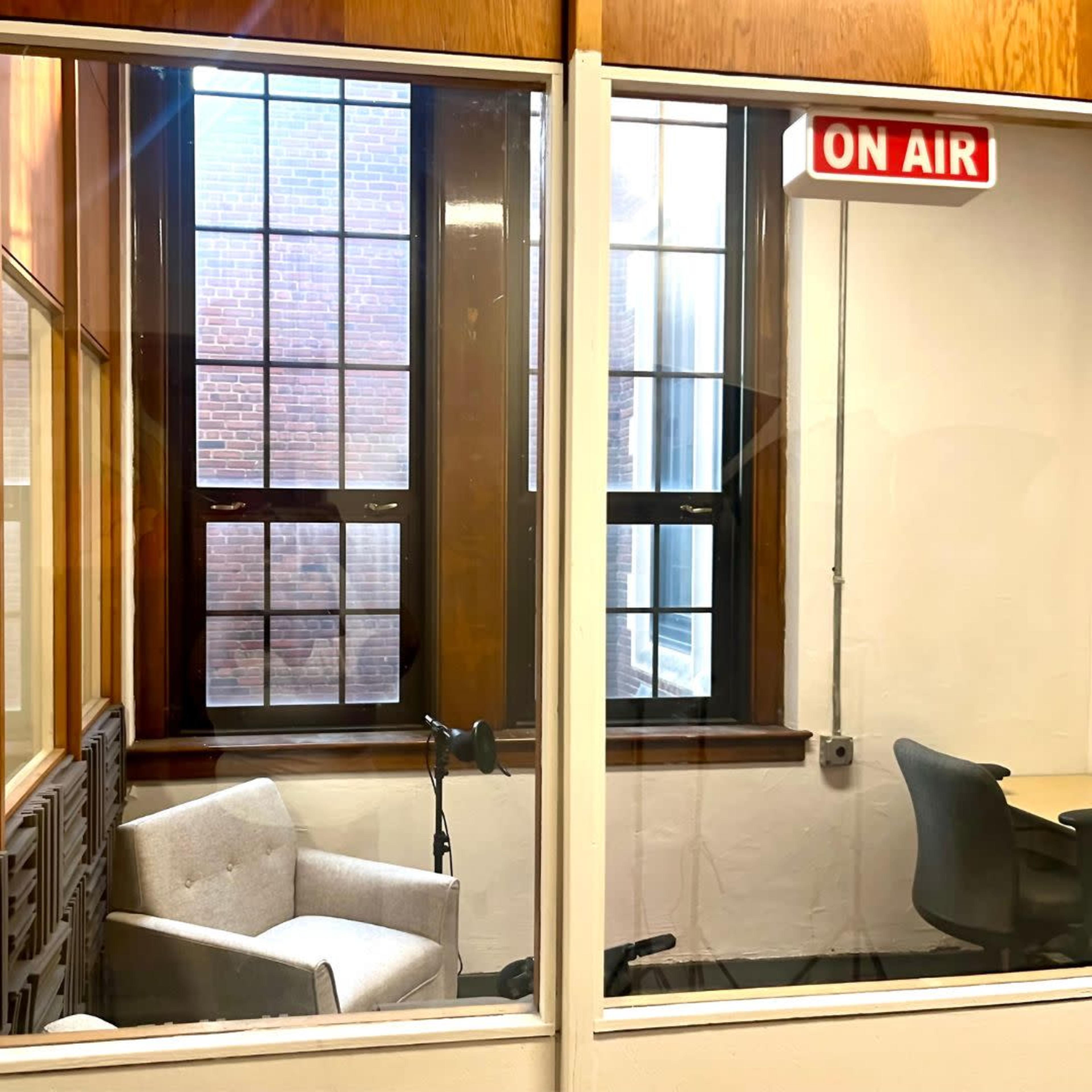 A well-lit room with a gray chair and a table visible through a glass window, featuring a red "ON AIR" sign above.