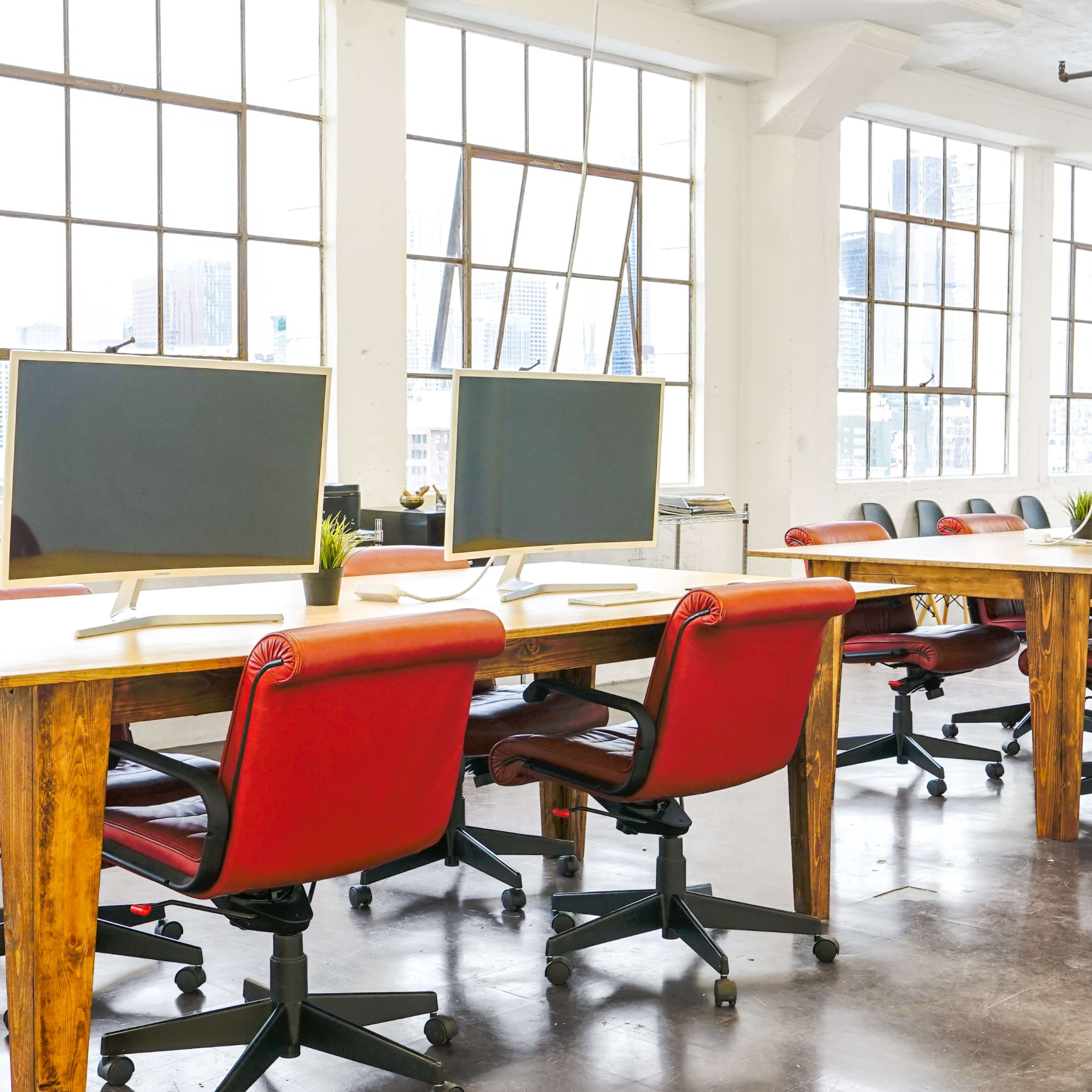A bright, open office space with wooden tables, red office chairs, and large windows offering a view of a city skyline.