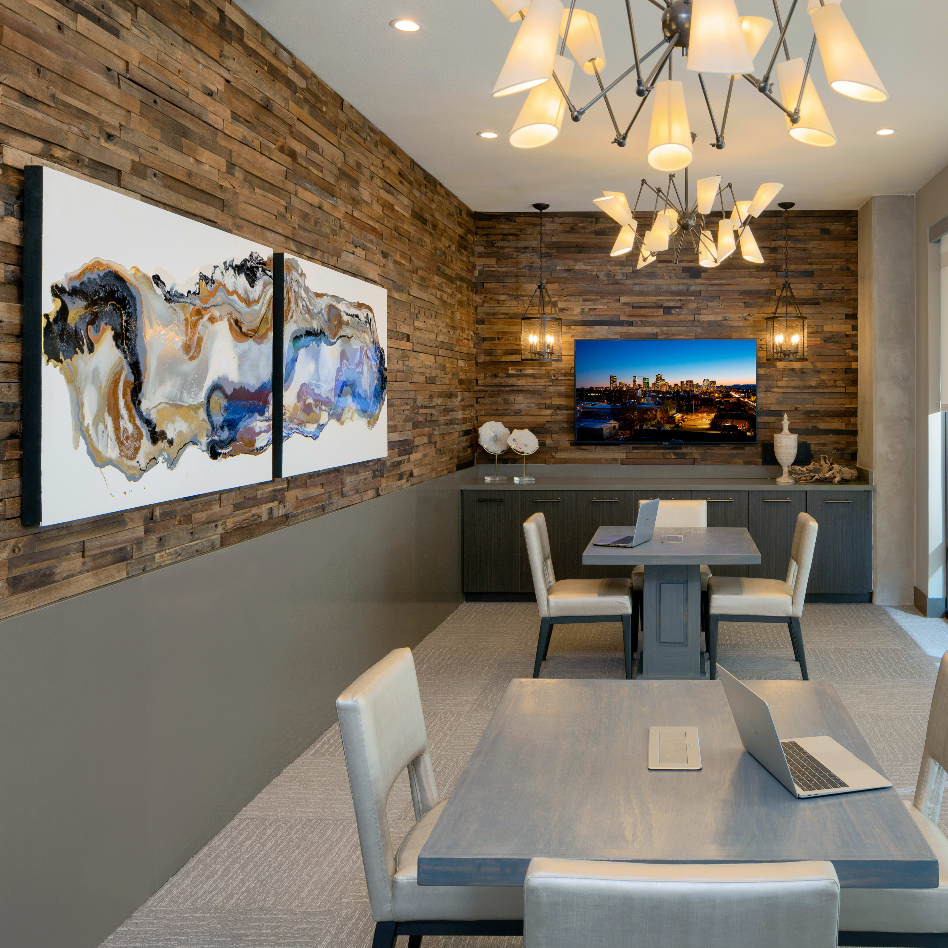 A modern conference room features a wooden accent wall, contemporary lighting, two artworks, a television, and four tables with chairs.