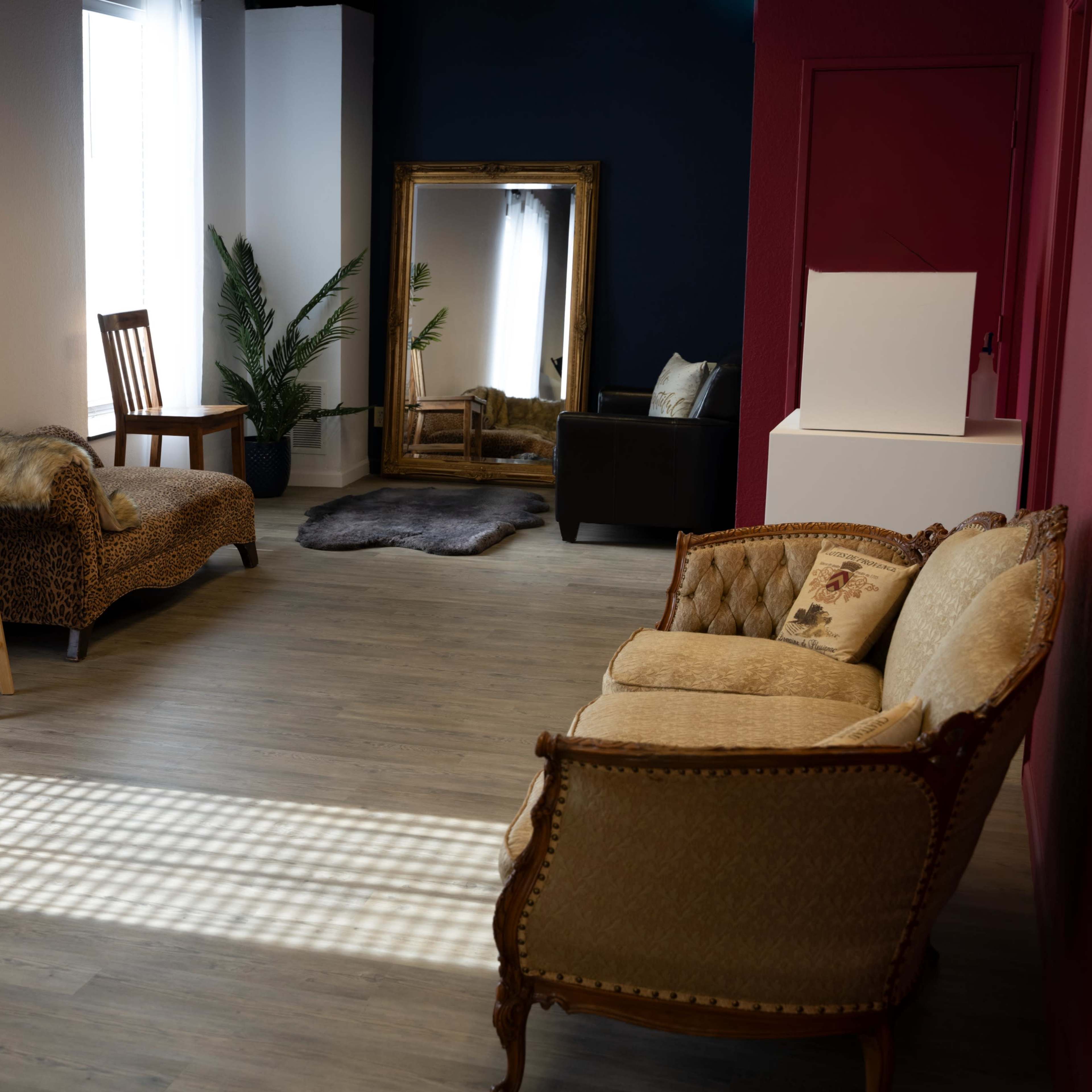 A spacious room with various seating arrangements, including a vintage couch, a wooden stool, and a chaise lounge, along with a large mirror and a potted plant.