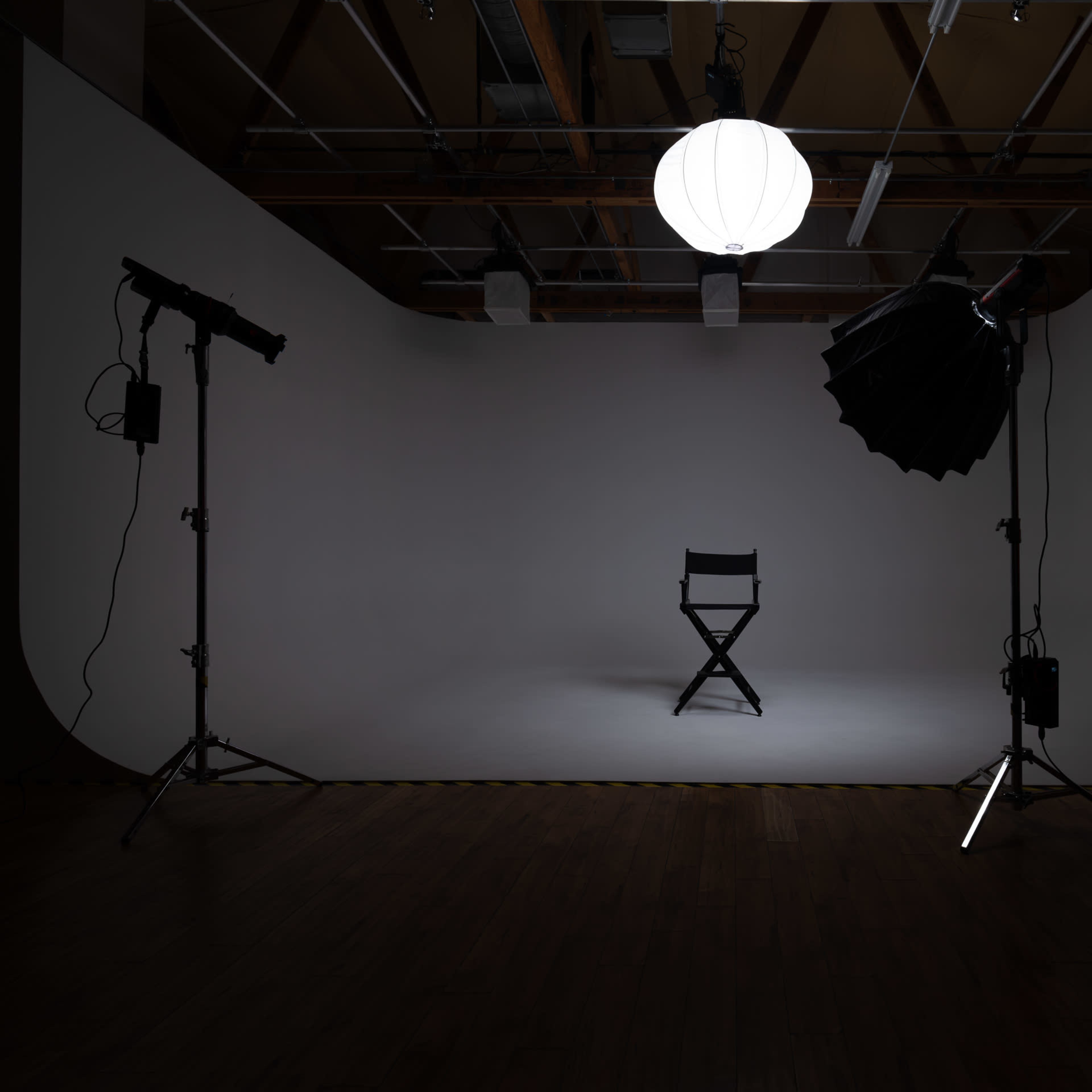 An empty photography studio with a single director's chair positioned under a large hanging light, flanked by studio lights on tripods.