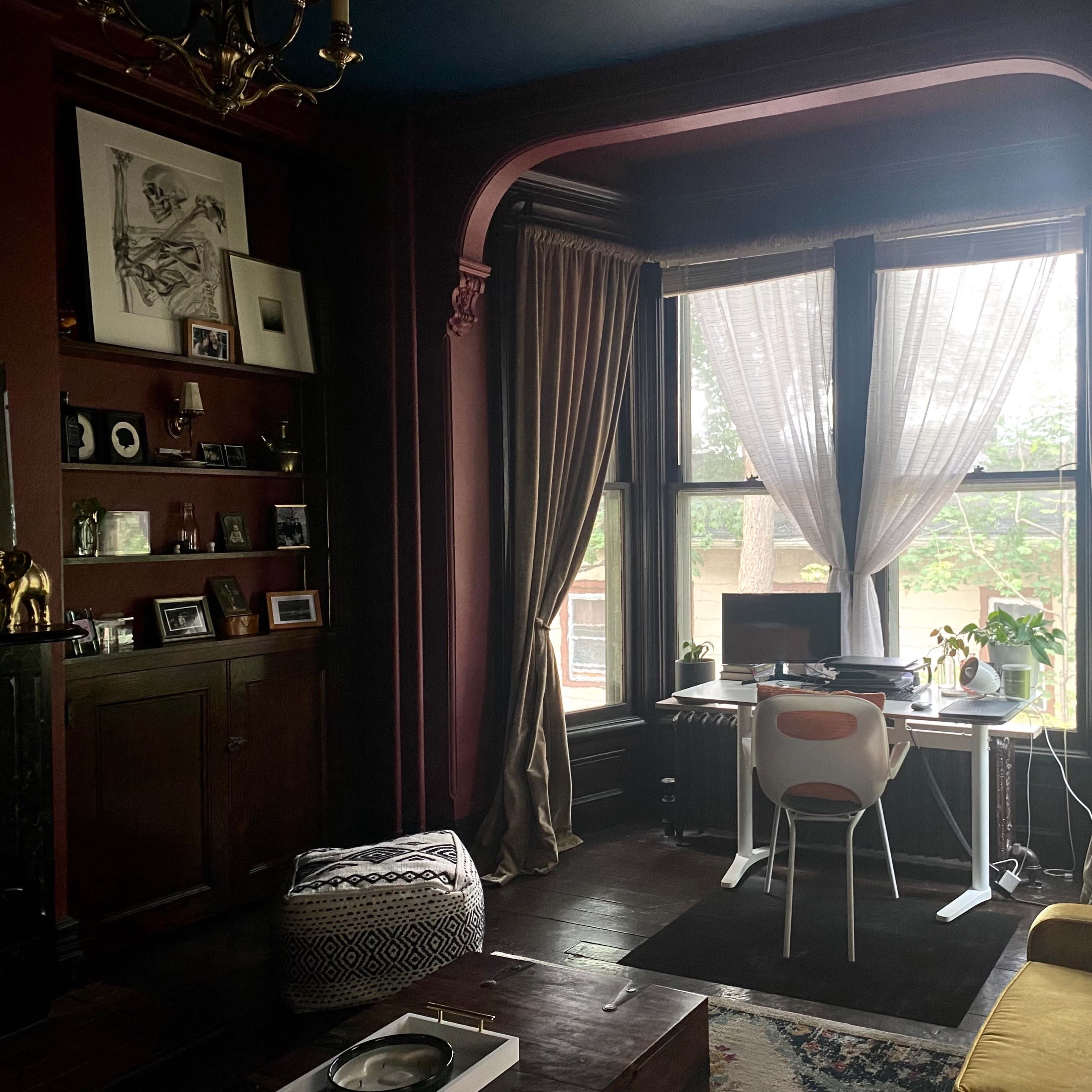 A dimly lit room features a desk by a large window, framed by drapes, with shelves and decorative items lining the walls.