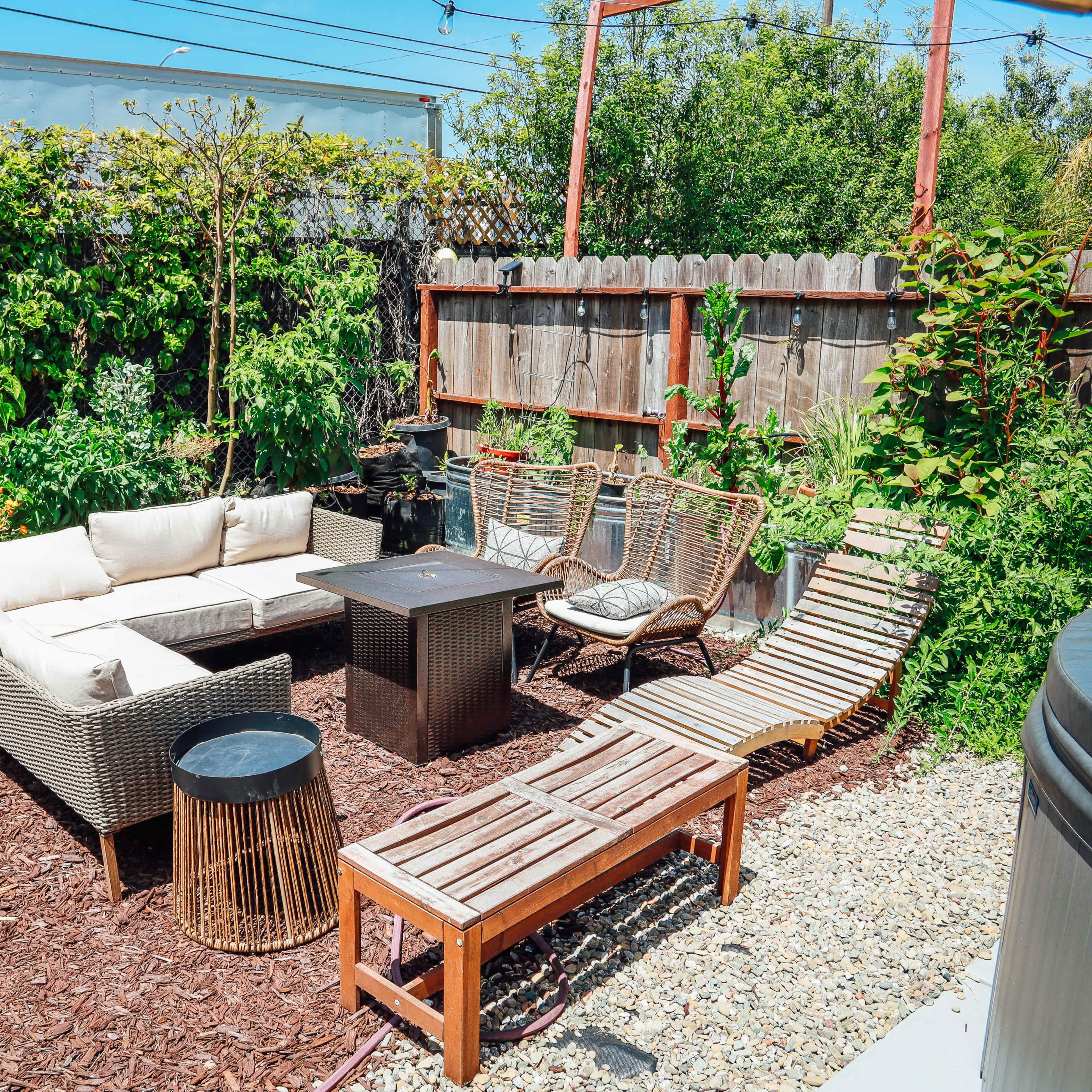 A cozy outdoor seating area features a sectional sofa, a coffee table, and lounge chairs surrounded by greenery and mulch.