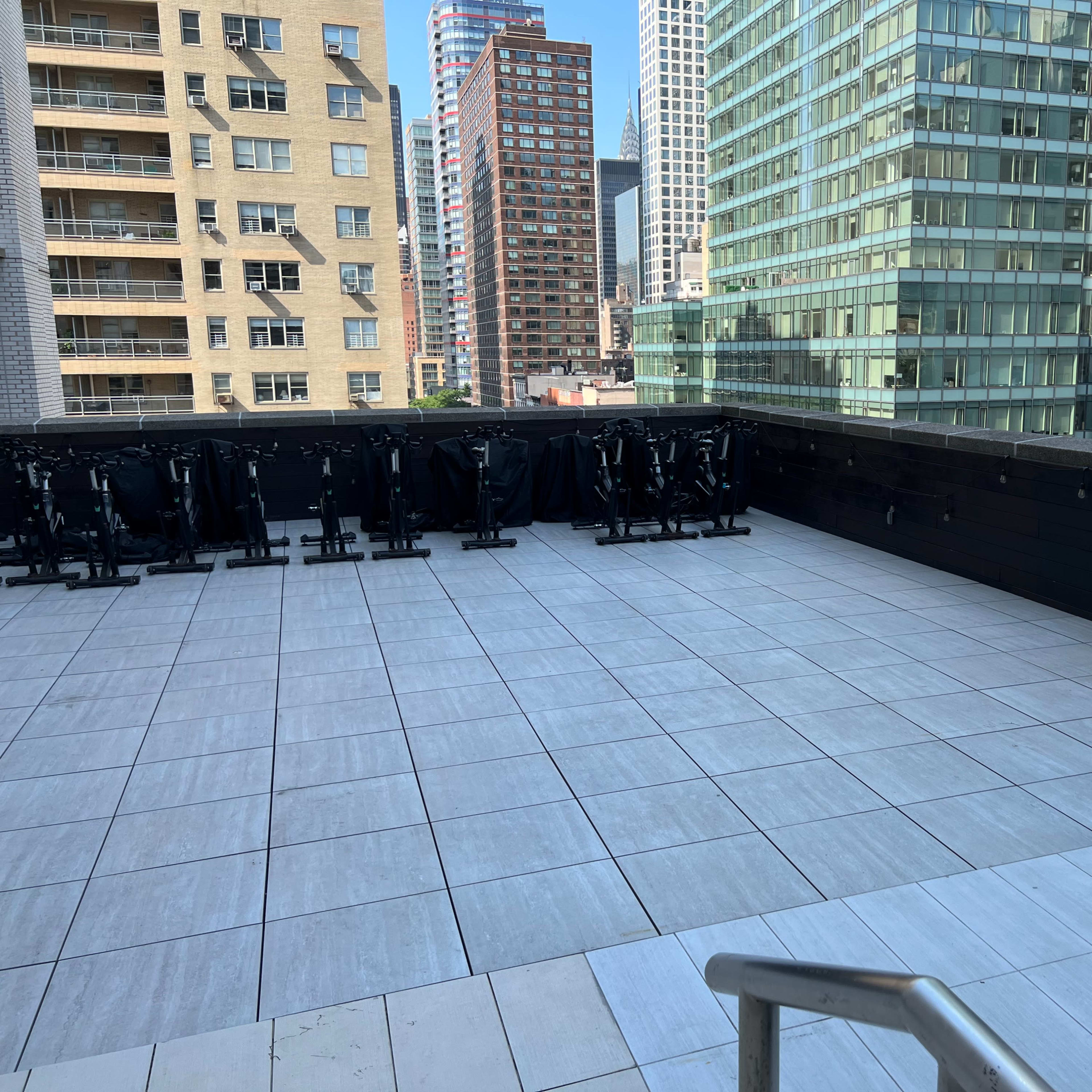 A partially empty rooftop terrace with tile flooring and stacked chairs against a railing, surrounded by tall buildings.