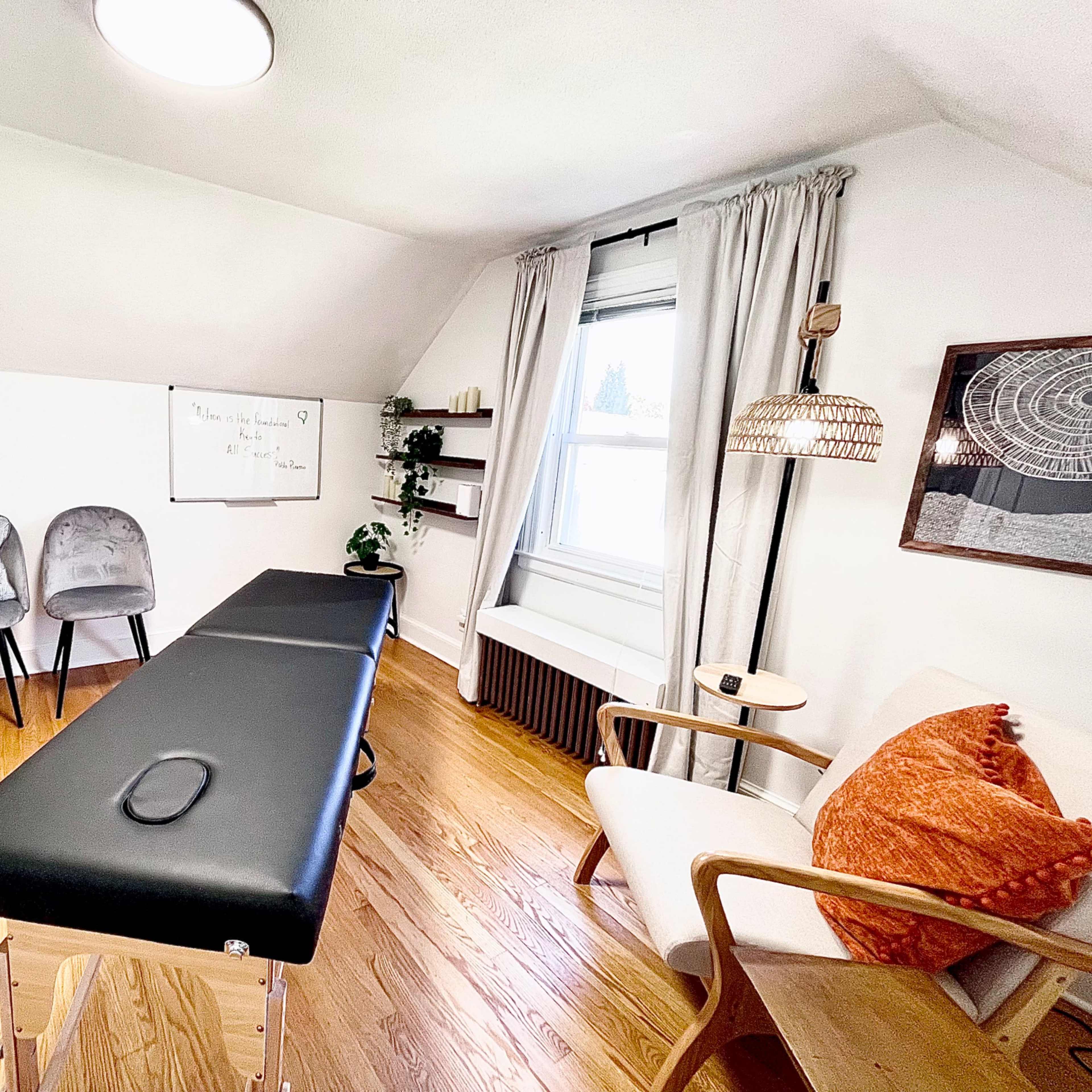 A compact treatment room with a therapy table, minimalist seating, and a window dressed with light curtains.