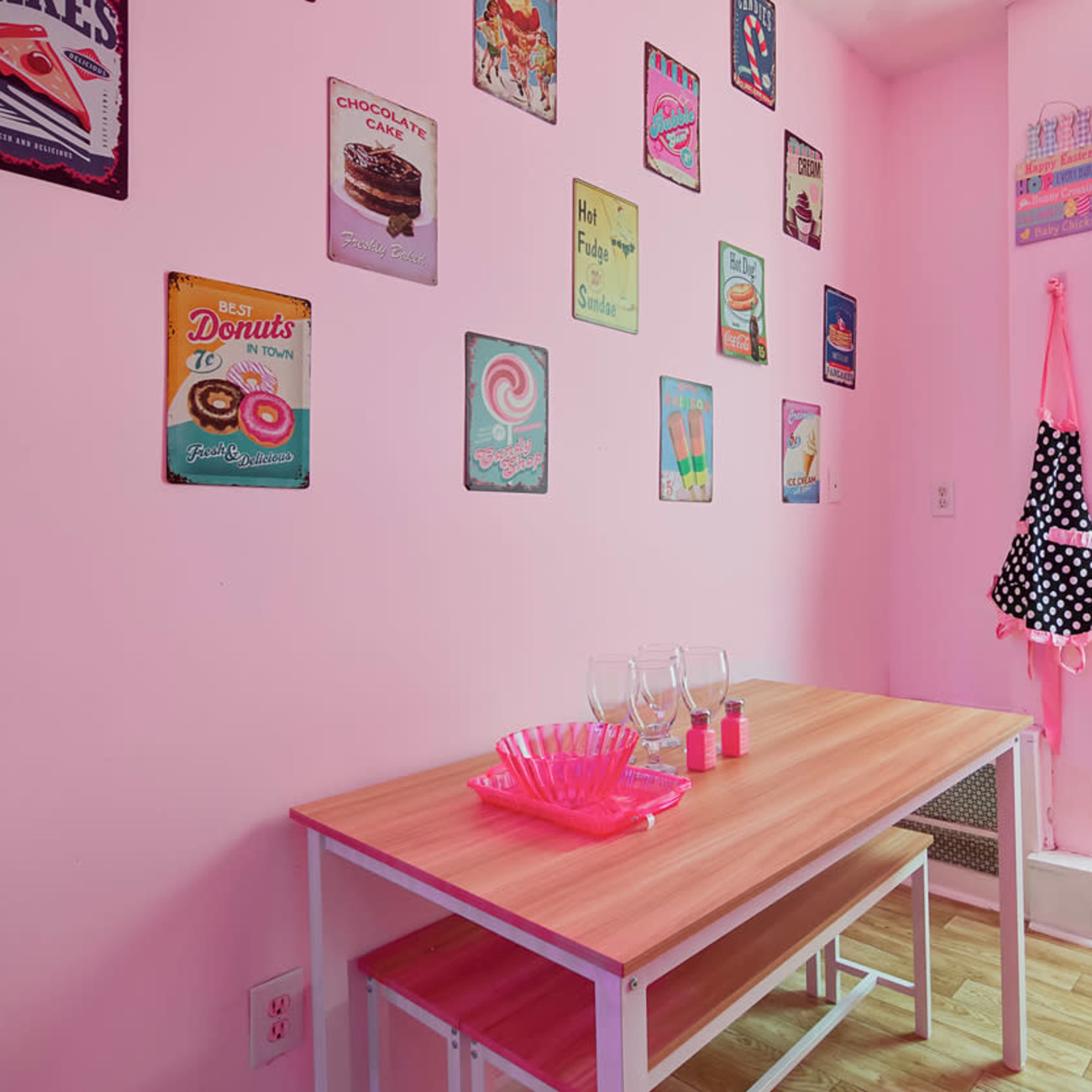 A small dining area features a wooden table with two benches, surrounded by pink walls adorned with vintage food-themed metal signs.