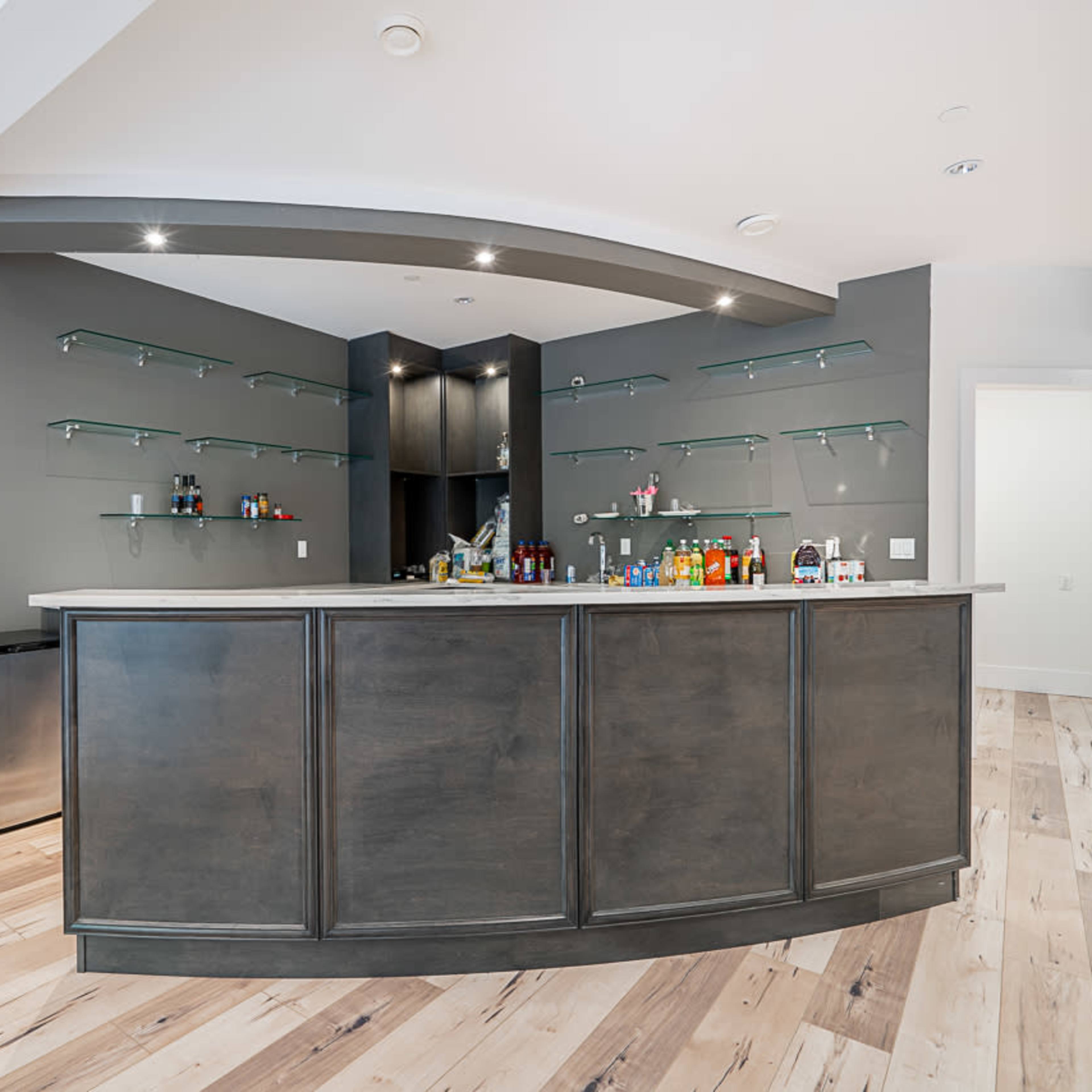 A modern home bar with a curved wooden countertop, mounted shelves displaying various bottles, and a minimalist design in a room with light wood flooring.