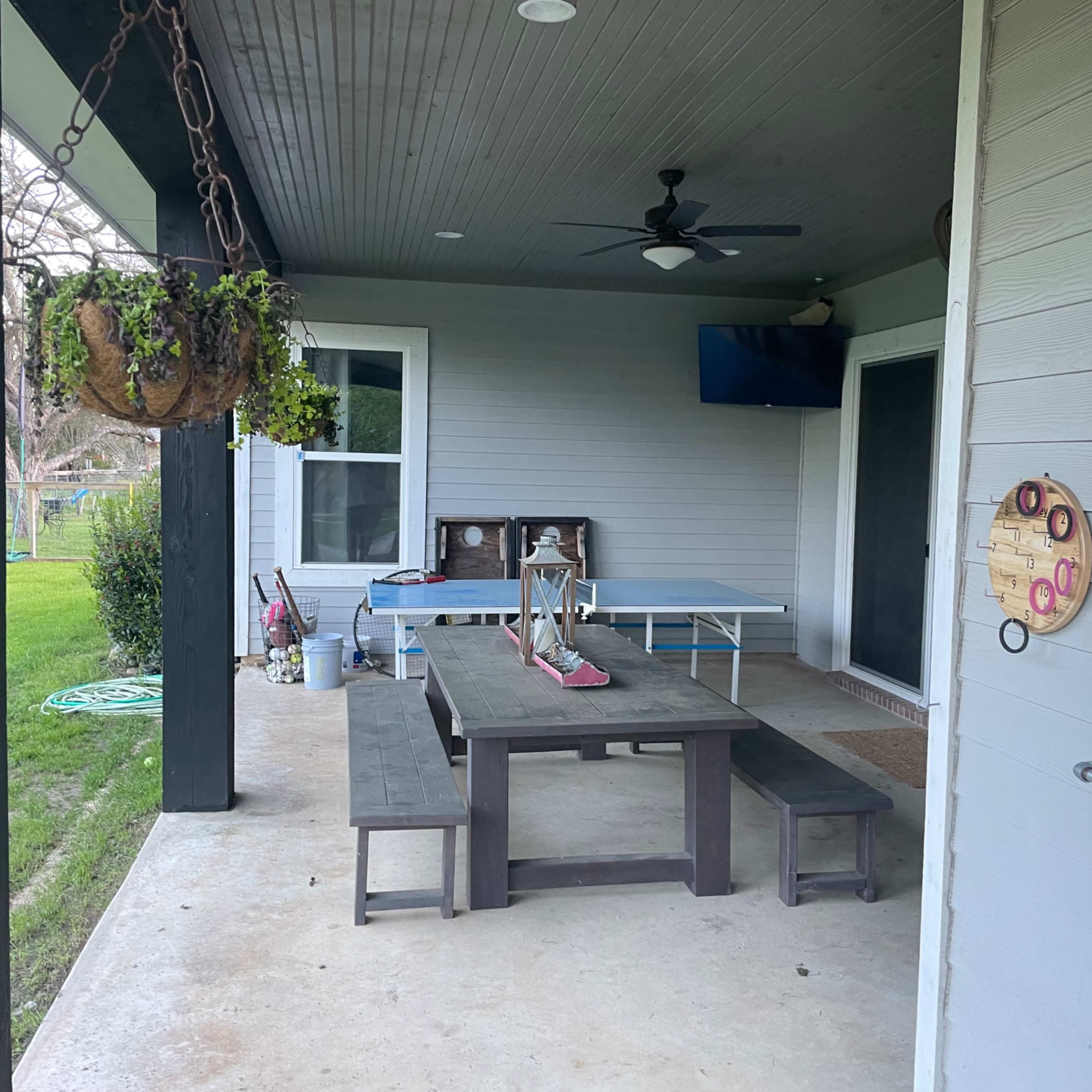 A covered patio area with a wooden table and benches, a ceiling fan, and a television mounted on the wall.