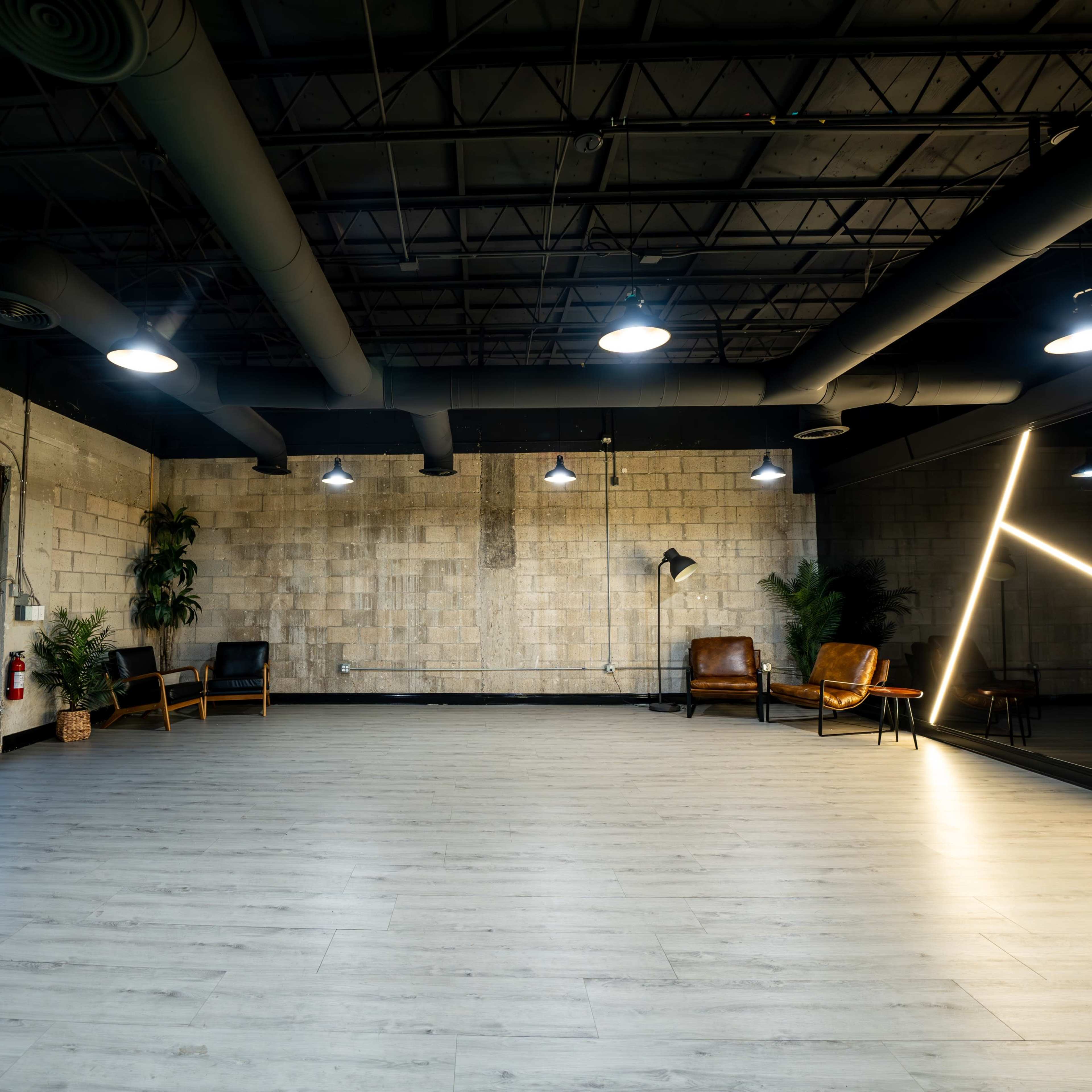 An empty studio space with concrete walls, wooden flooring, and two chairs arranged near a large mirror, illuminated by overhead lights.
