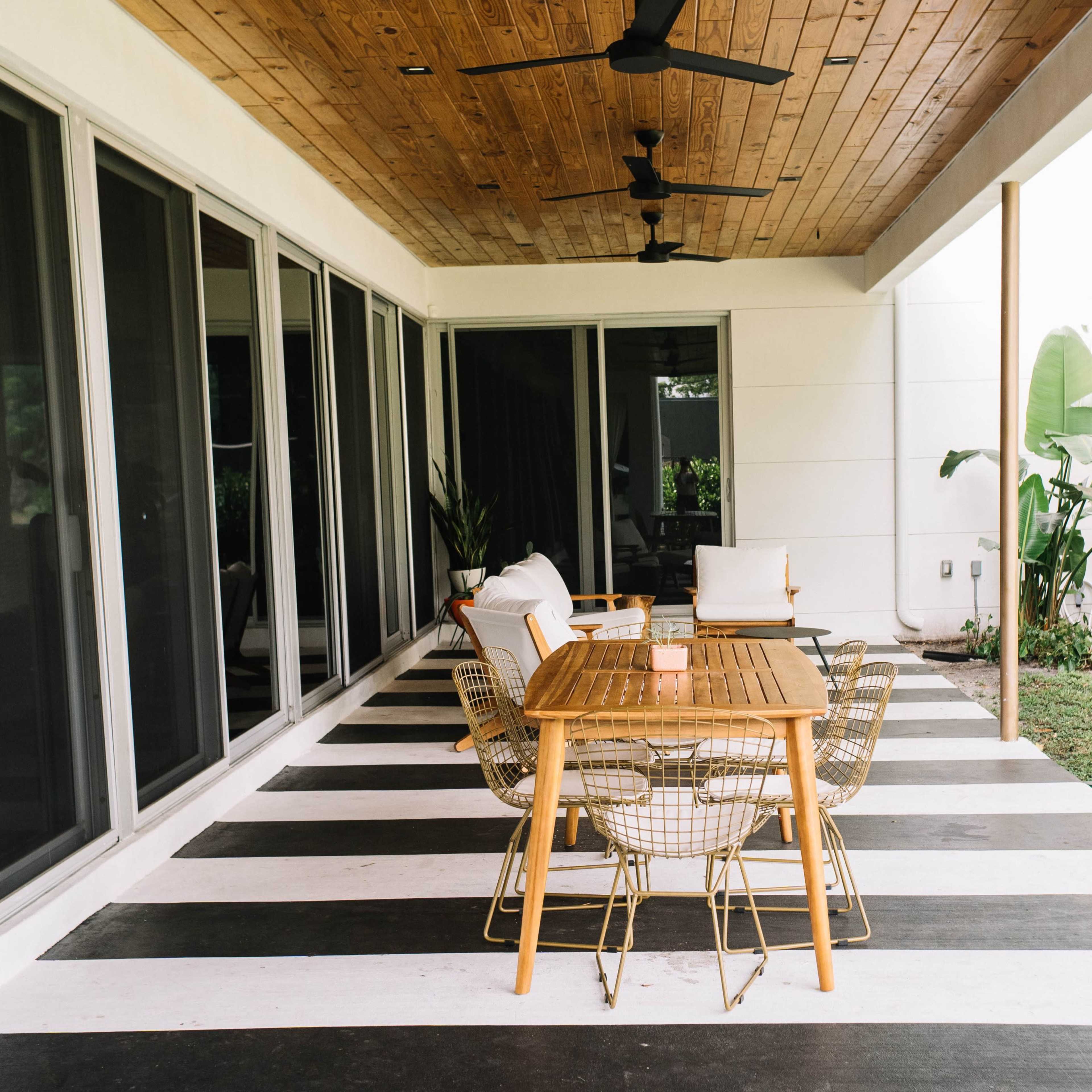 A covered outdoor patio with a wooden dining table and four wire chairs on a black and white striped floor.