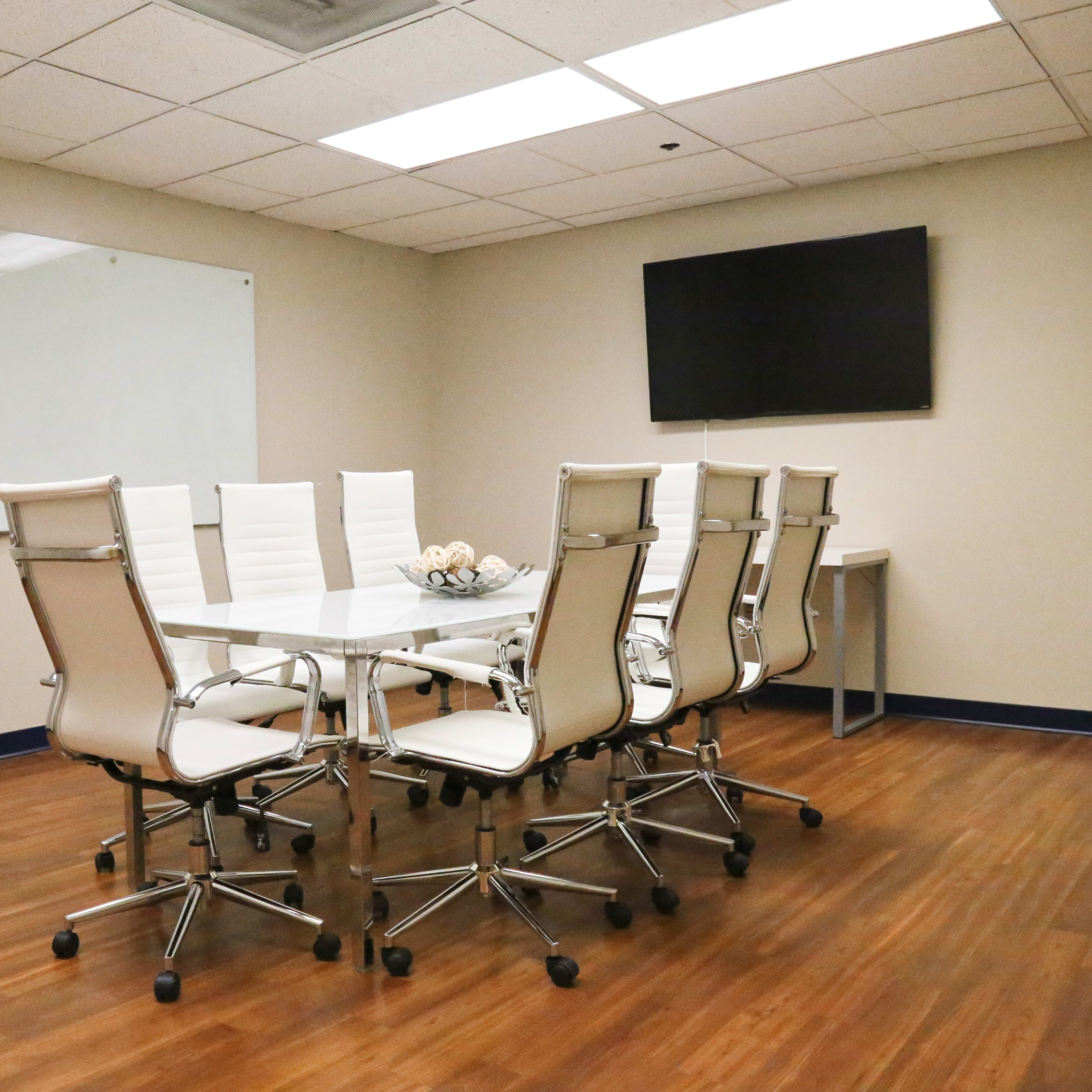 A modern conference room features a large glass table surrounded by eight white swivel chairs, with a wall-mounted TV and a whiteboard visible in the background.