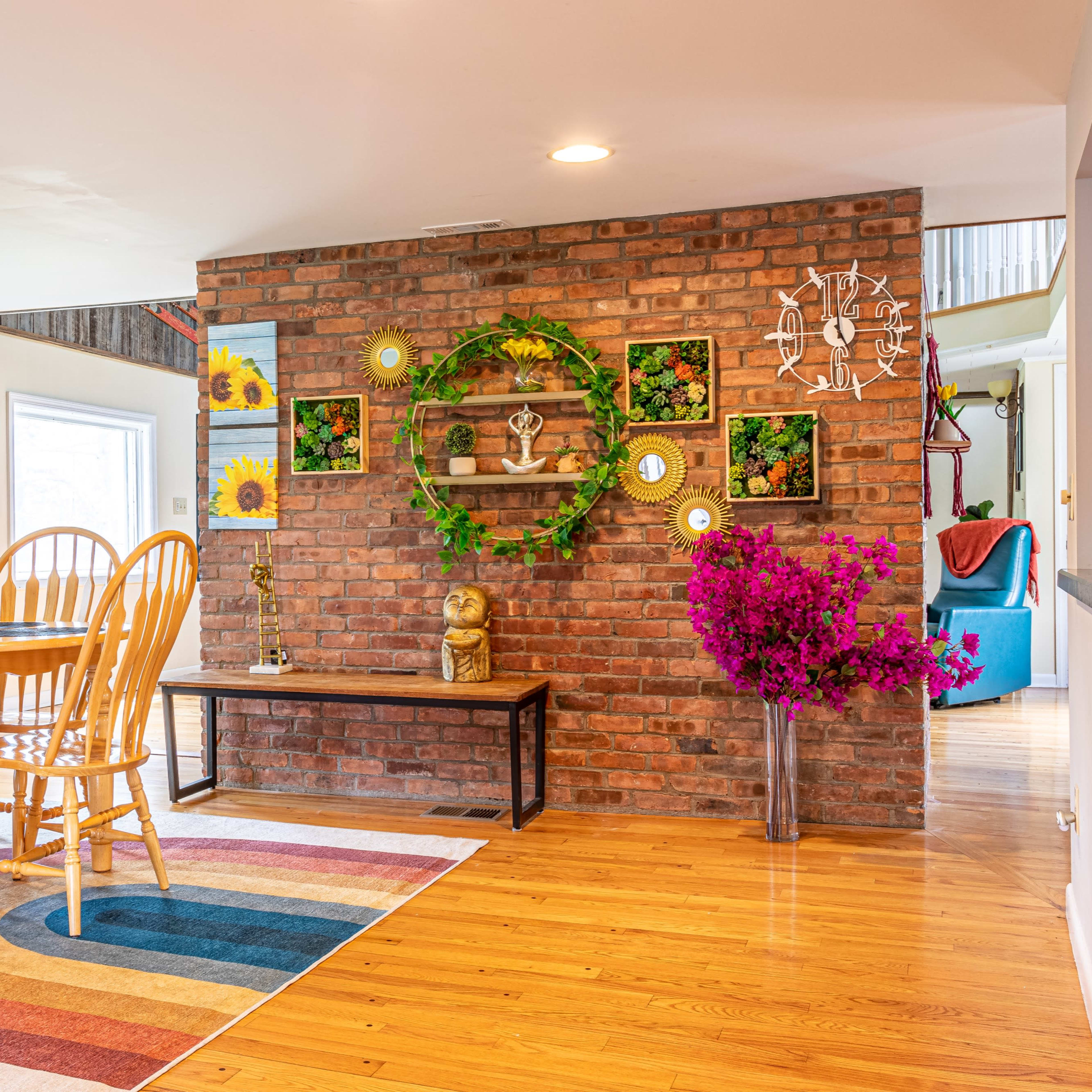 A dining area featuring a circular wooden table surrounded by six wooden chairs, a brick accent wall adorned with decorative items, and a colorful area rug.